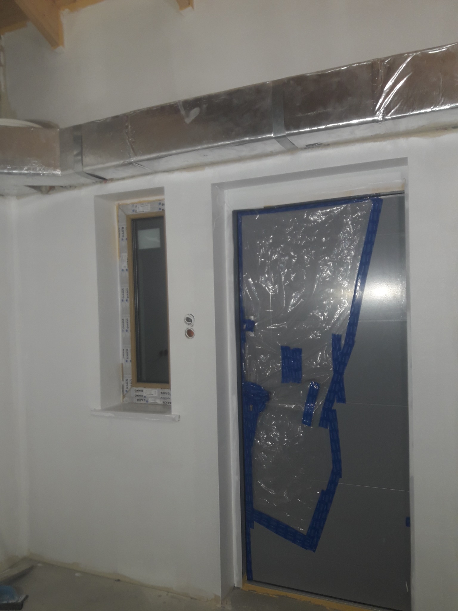 Świeżo osadzone okno PCV i zabezpieczone folią drzwi w surowym wnętrzu. Widoczny fragment instalacji wentylacyjnej pod sufitem. Ściany przygotowane do malowania.