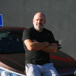 DUET Damian Dobiecki Ośrodek Szkolenia Kierowców Galanteria ZUZA sklep internetowy - Instruktor oparty o auto z oznaczeniem 'L' i napisem 'DUET', skrzyżowane ręce, poważny wyraz twarzy, szare tło. Nietypowe auto szkoły jazdy.