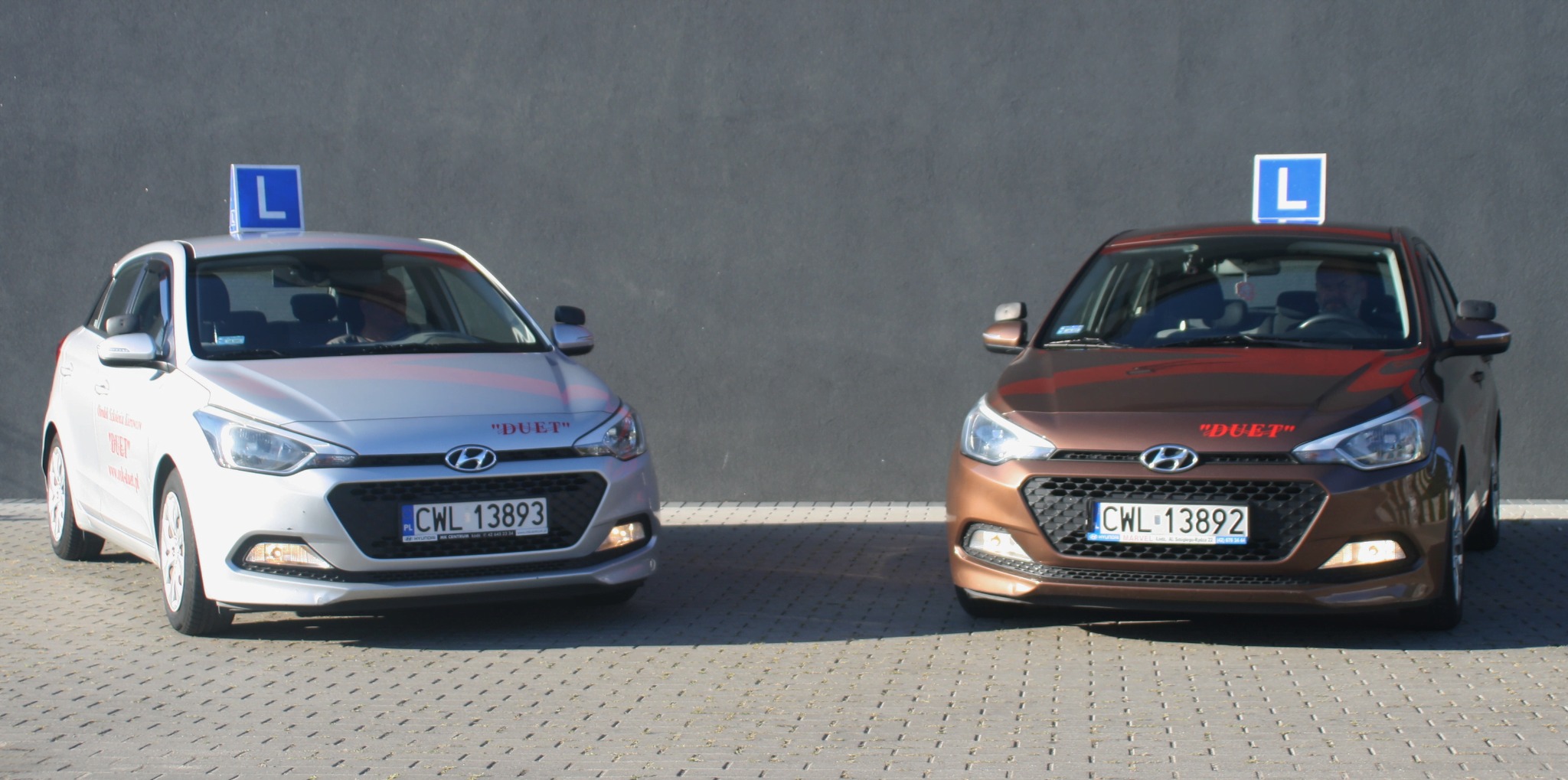 Dwa samochody nauki jazdy, srebrny i brązowy, z oznaczeniem 'L' na dachu, zaparkowane na brukowanym placu przed szarą ścianą. Widoczne numery rejestracyjne.