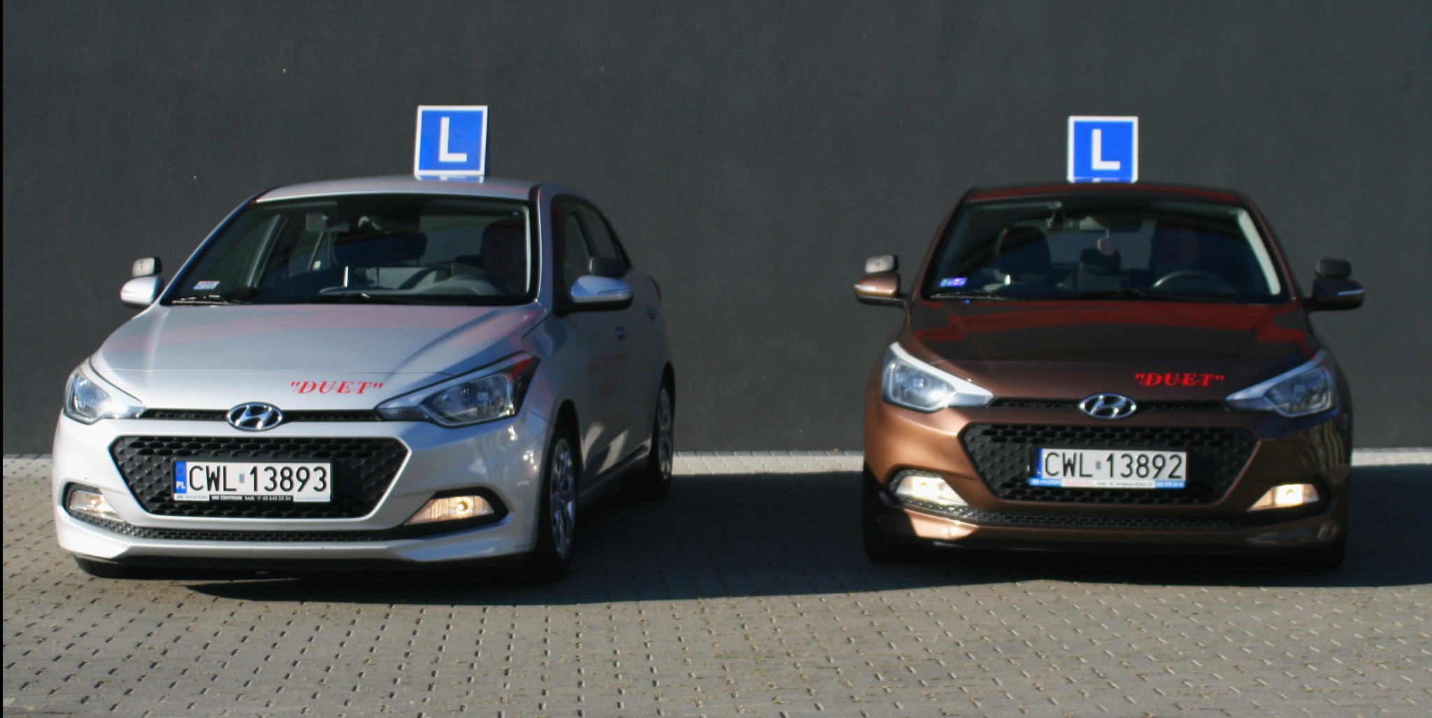 Dwa samochody Hyundai i20 z literą 'L' na dachu, srebrny i brązowy, zaparkowane przed szarą ścianą. Tablice rejestracyjne CWL. Napis 'DUET' nad logo.