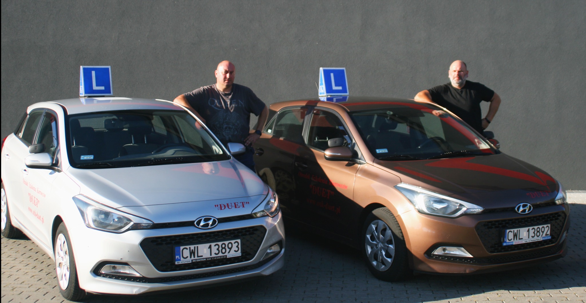 Dwa samochody z literą 'L' na dachu, używane w szkole jazdy 'DUET' w Wichrowicach, zaparkowane obok siebie. Instruktorzy opierają się o maski pojazdów.