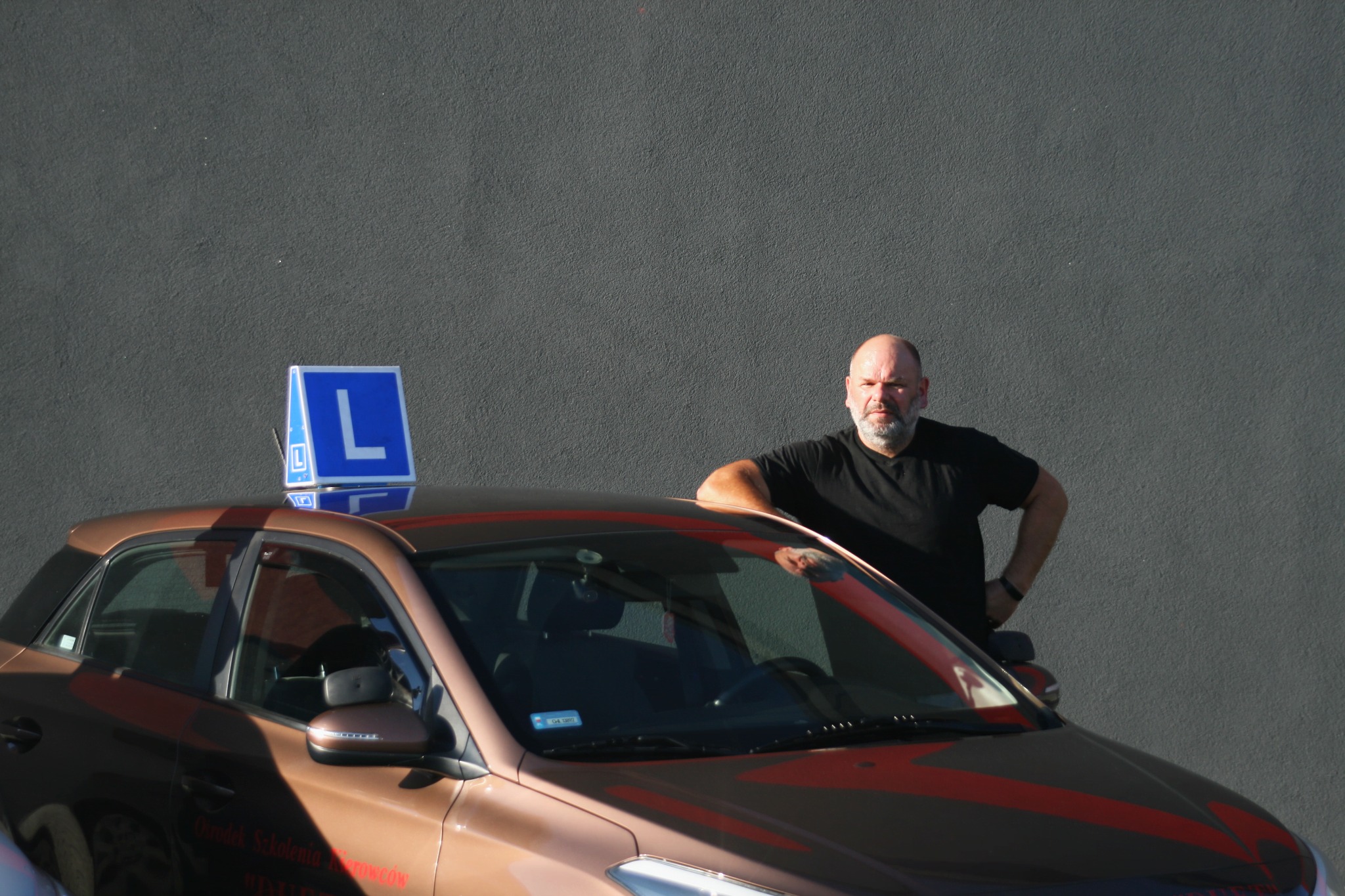 Instruktor oparty o brązowy samochód nauki jazdy z niebieską literą L na dachu, na tle szarej ściany. Widoczny fragment tablicy rejestracyjnej i naklejki szkoły.