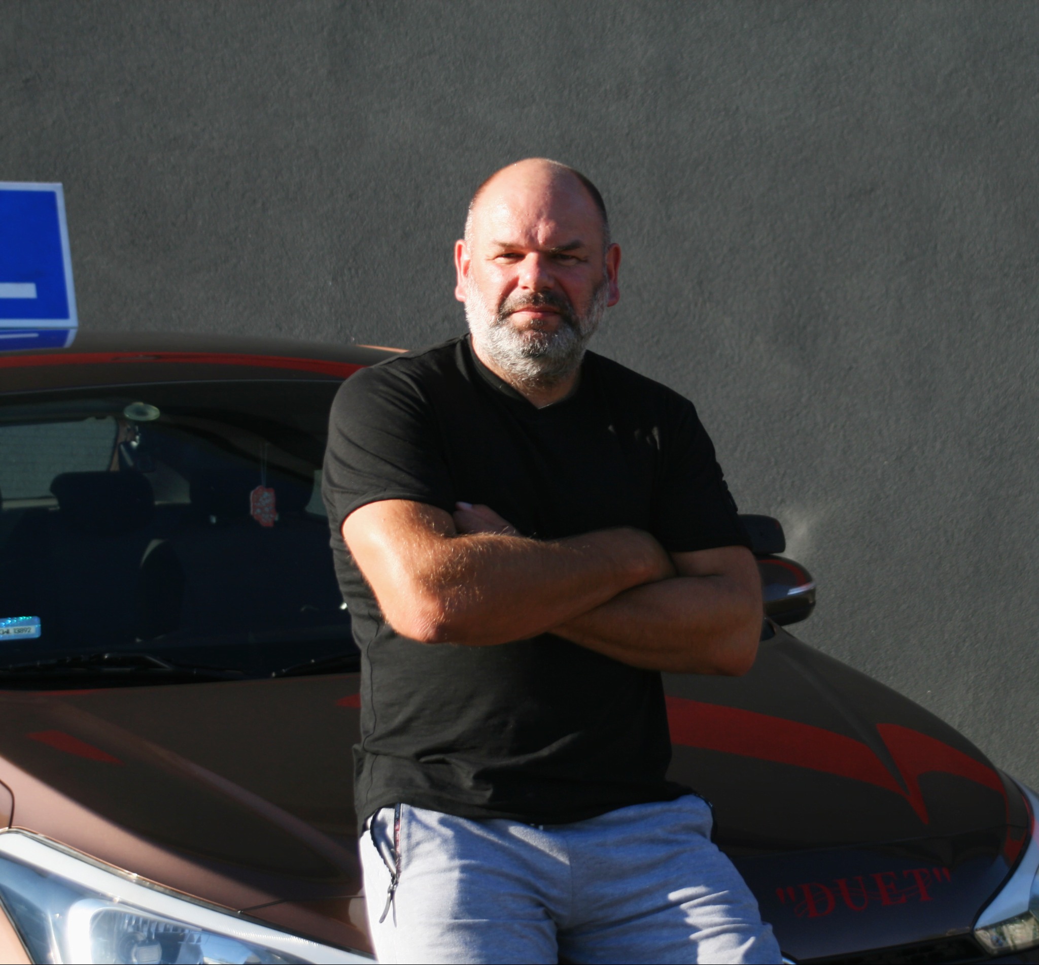 Instruktor oparty o auto z oznaczeniem 'L' i napisem 'DUET', skrzyżowane ręce, poważny wyraz twarzy, szare tło. Nietypowe auto szkoły jazdy.