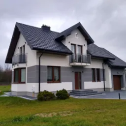 Dwukondygnacyjny dom z ciemnoszarą dachówką, balkonem z balustradą ze szkła i szarymi akcentami na elewacji, otoczony trawnikiem i kostką brukową.