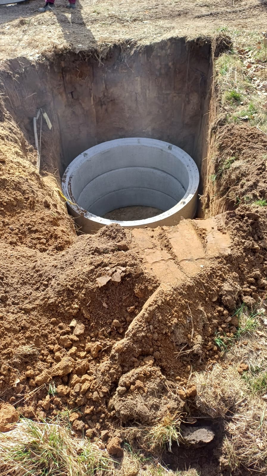 Betonowy krąg w wykopie ziemnym, prawdopodobnie element szamba lub studni. Widoczne ślady pracy ziemnej i przygotowanie do dalszych etapów instalacji.