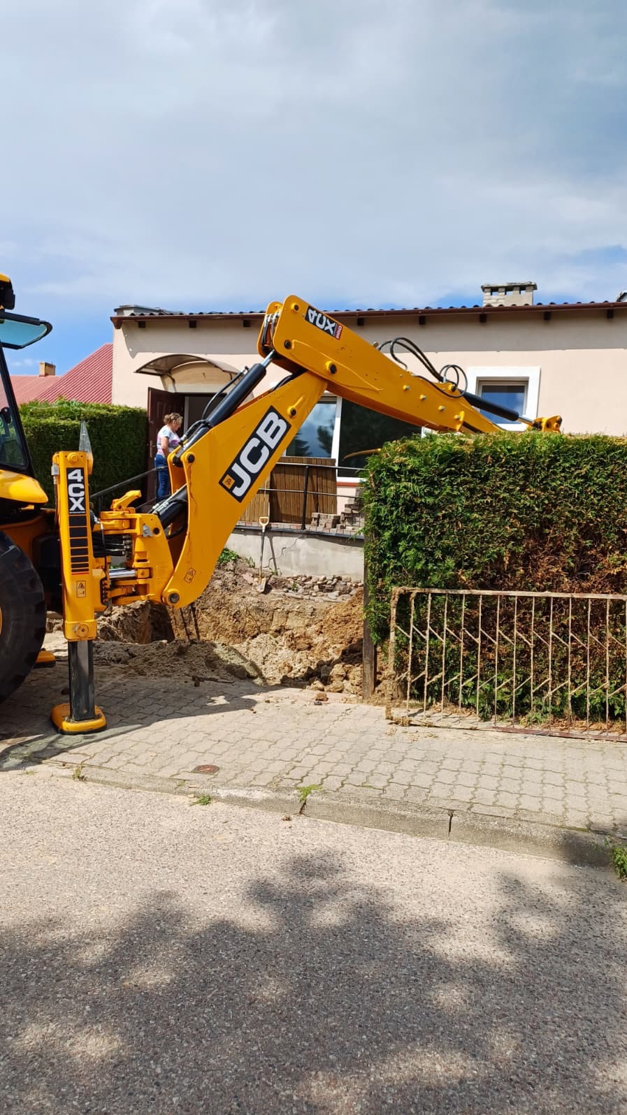 Żółta koparka JCB podczas prac ziemnych przy posesji, widoczny wykop w ziemi, fragment ogrodzenia i osoba w tle. Praca w słoneczny dzień.