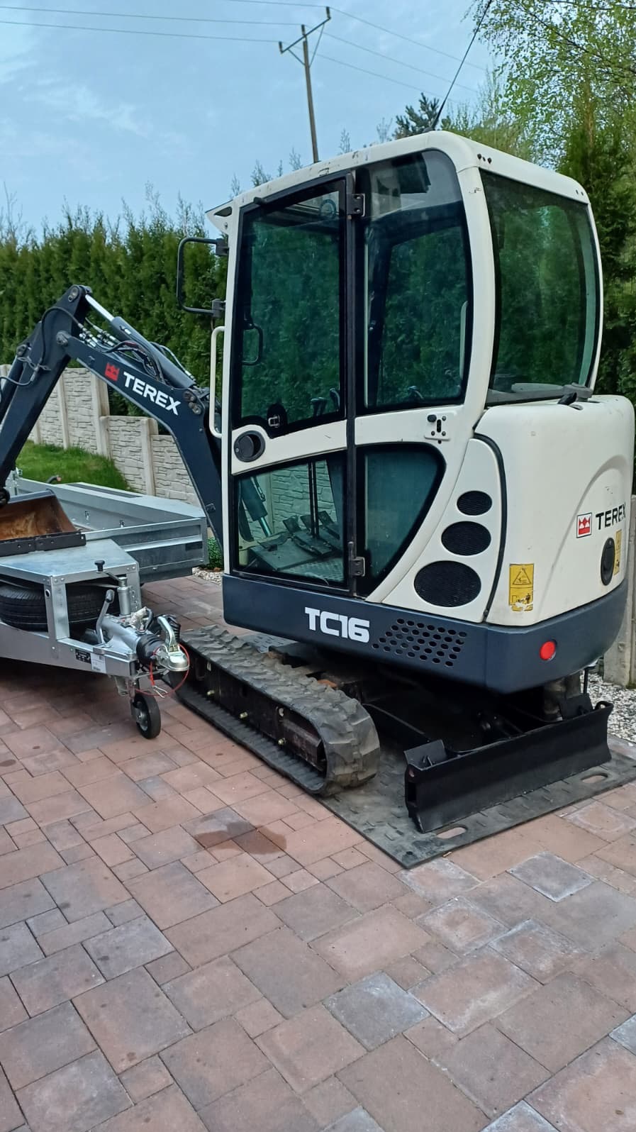 Kompaktowa minikoparka Terex TC16 na gąsienicach, z przyczepą, zaparkowana na kostce brukowej. Widoczne logo Terex i oznaczenie modelu TC16.