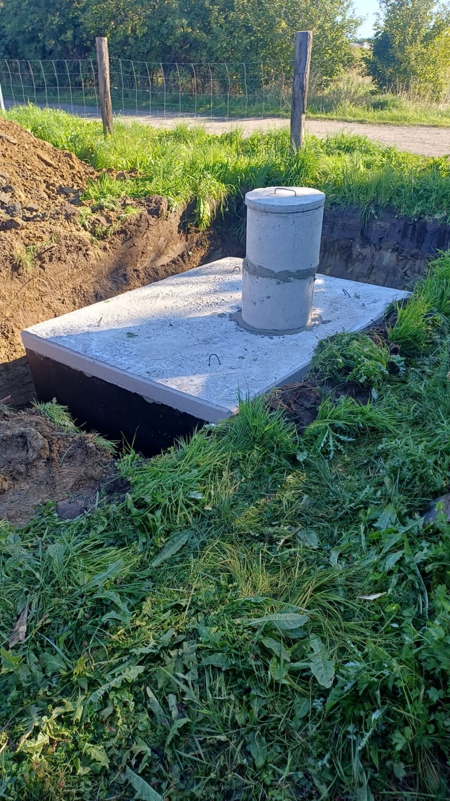 Betonowy zbiornik z włazem w wykopie, otoczony zielenią. Widoczne ślady ziemi i trawy, co sugeruje świeżo zakończone prace instalacyjne. Ujęcie z poziomu gruntu.