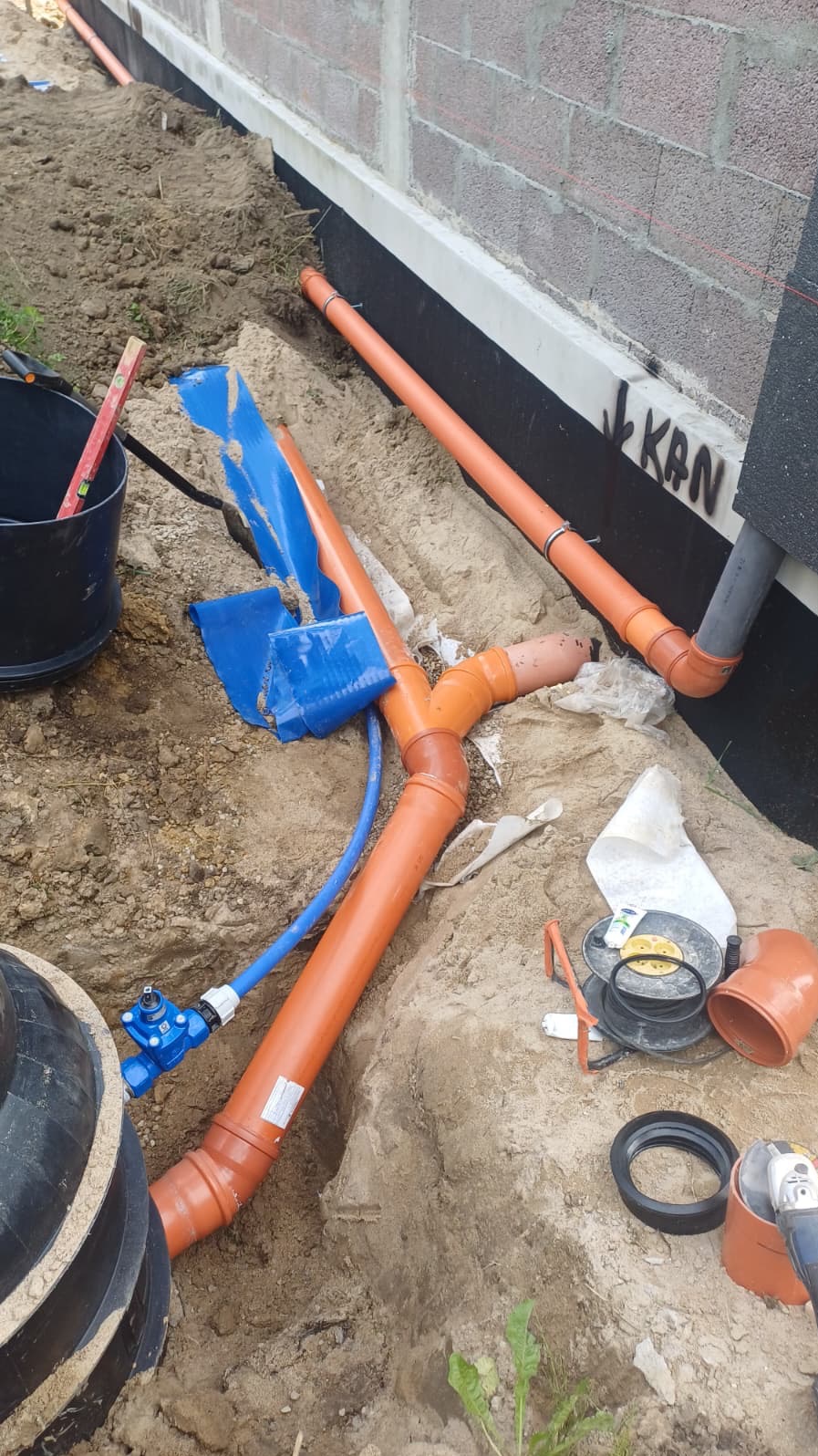 Pomarańczowe rury kanalizacyjne w wykopie przy fundamencie budynku, z widocznym niebieskim zaworem i elementami instalacji. Częściowo zasypane ziemią.