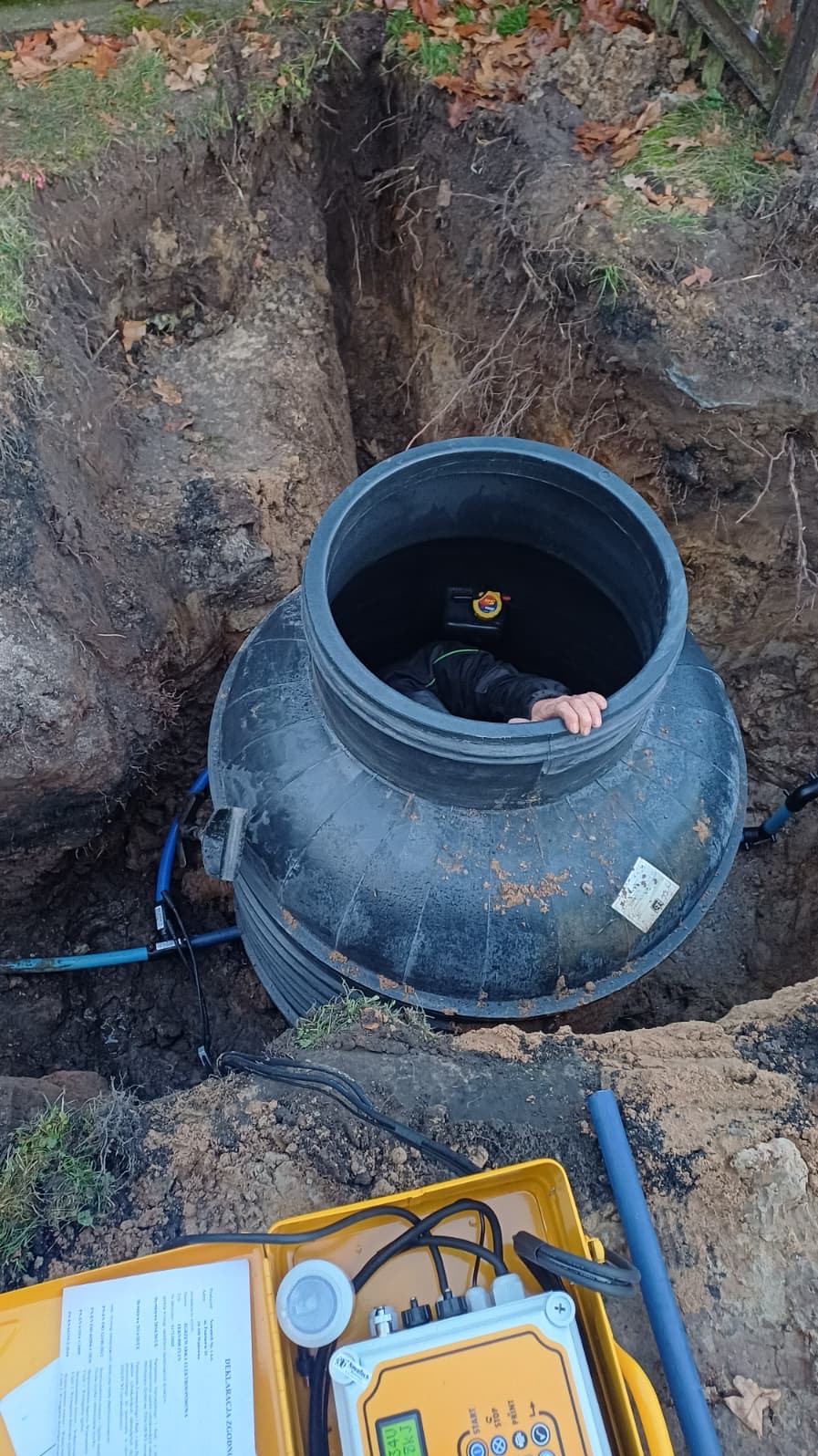 Montaż szamba: Czarna, plastikowa konstrukcja w wykopie, widoczna ręka pracownika wewnątrz. Żółta skrzynka z urządzeniem pomiarowym na pierwszym planie.
