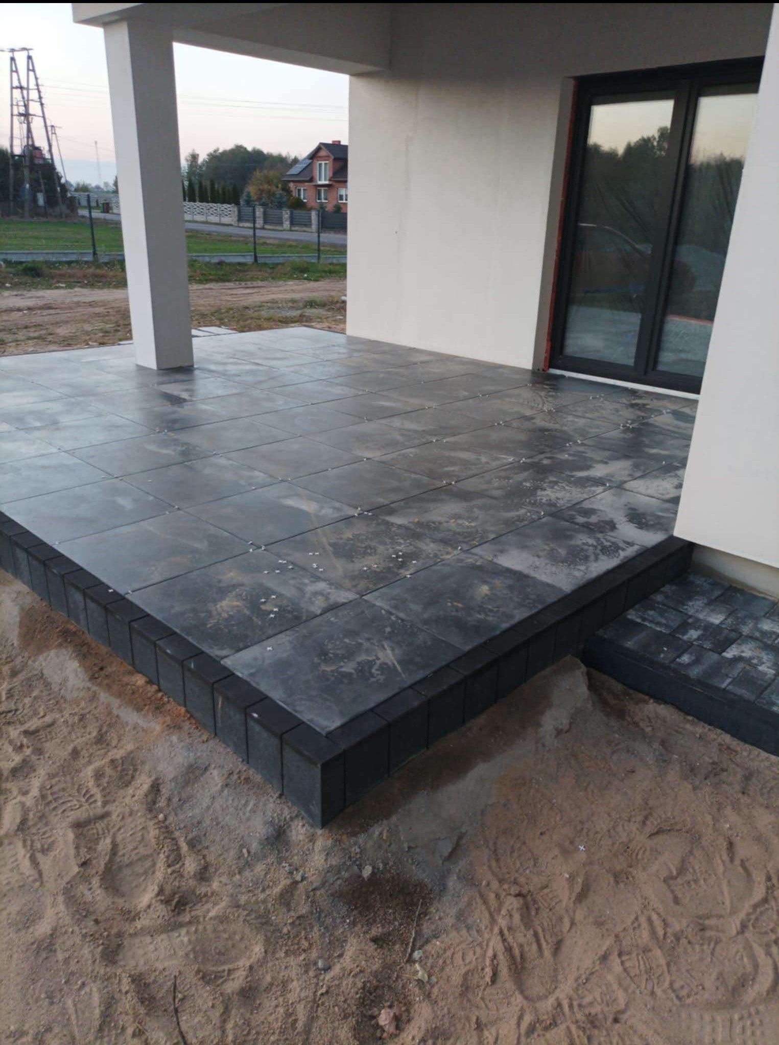 Nowoczesny taras z szarych płyt, obrzeża z ciemnej kostki. Widoczne ślady montażu, piasek. W tle dom z czarnymi oknami i słup podtrzymujący zadaszenie.