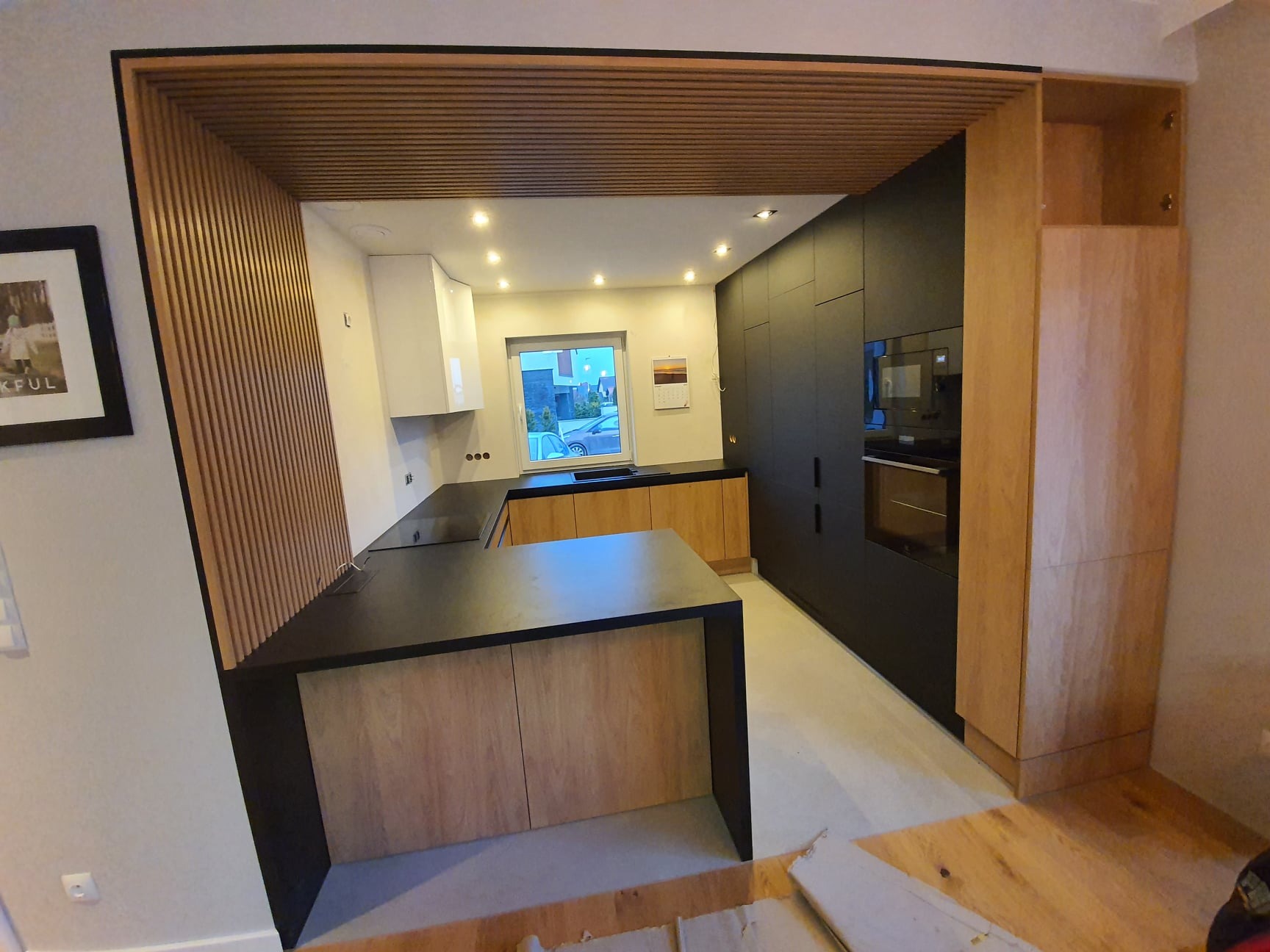 Nowoczesna kuchnia z wyspą i drewnianymi lamelami. Czarny blat kontrastuje z jasnym drewnem szafek. Widoczne zabudowane sprzęty i okno z widokiem na zewnątrz.