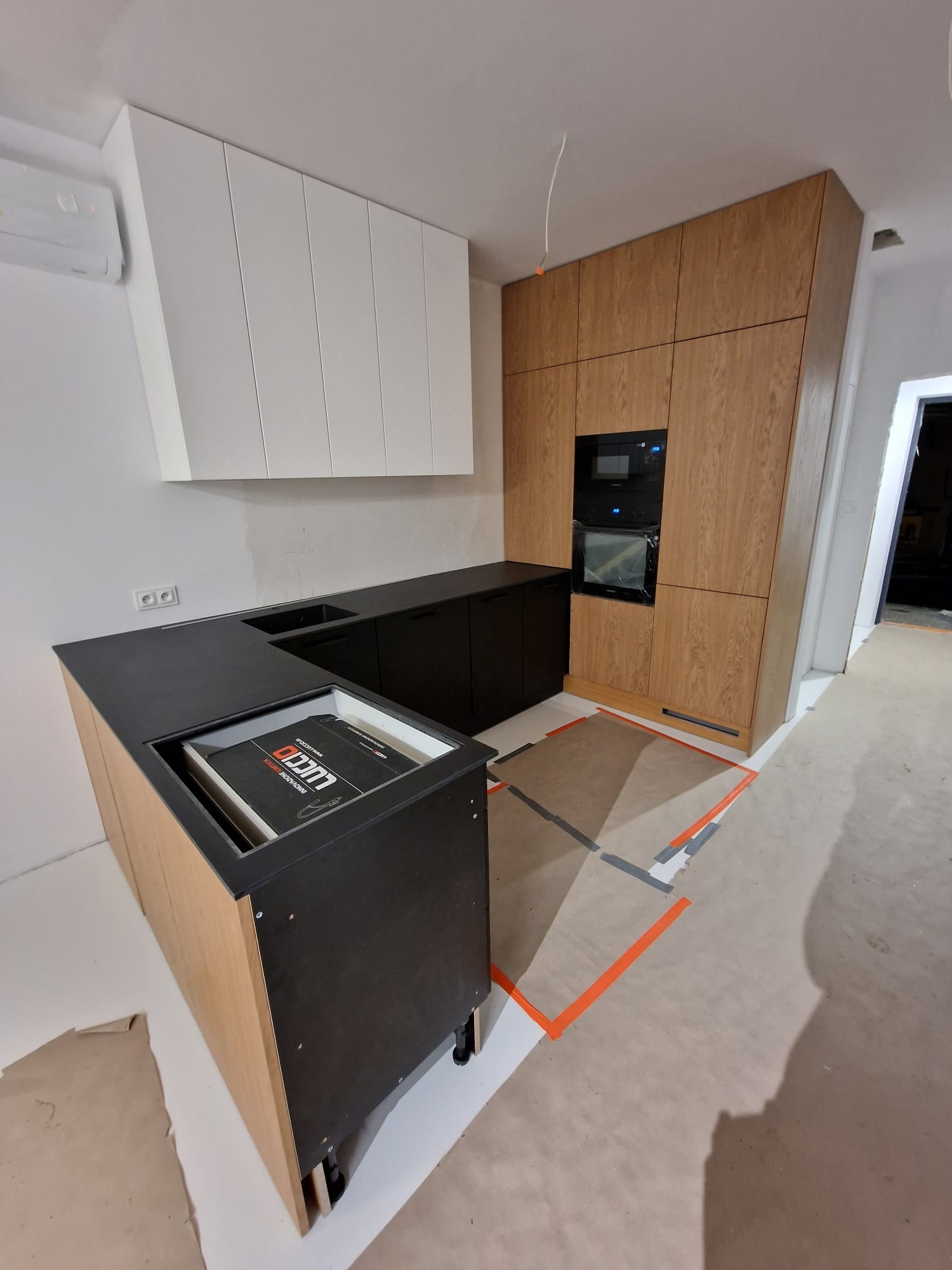 Częściowo zamontowana kuchnia na wymiar z czarnym blatem i drewnianymi frontami. Widoczne kartony z akcesoriami. Oświetlenie sufitowe. Podłoga zabezpieczona folią.