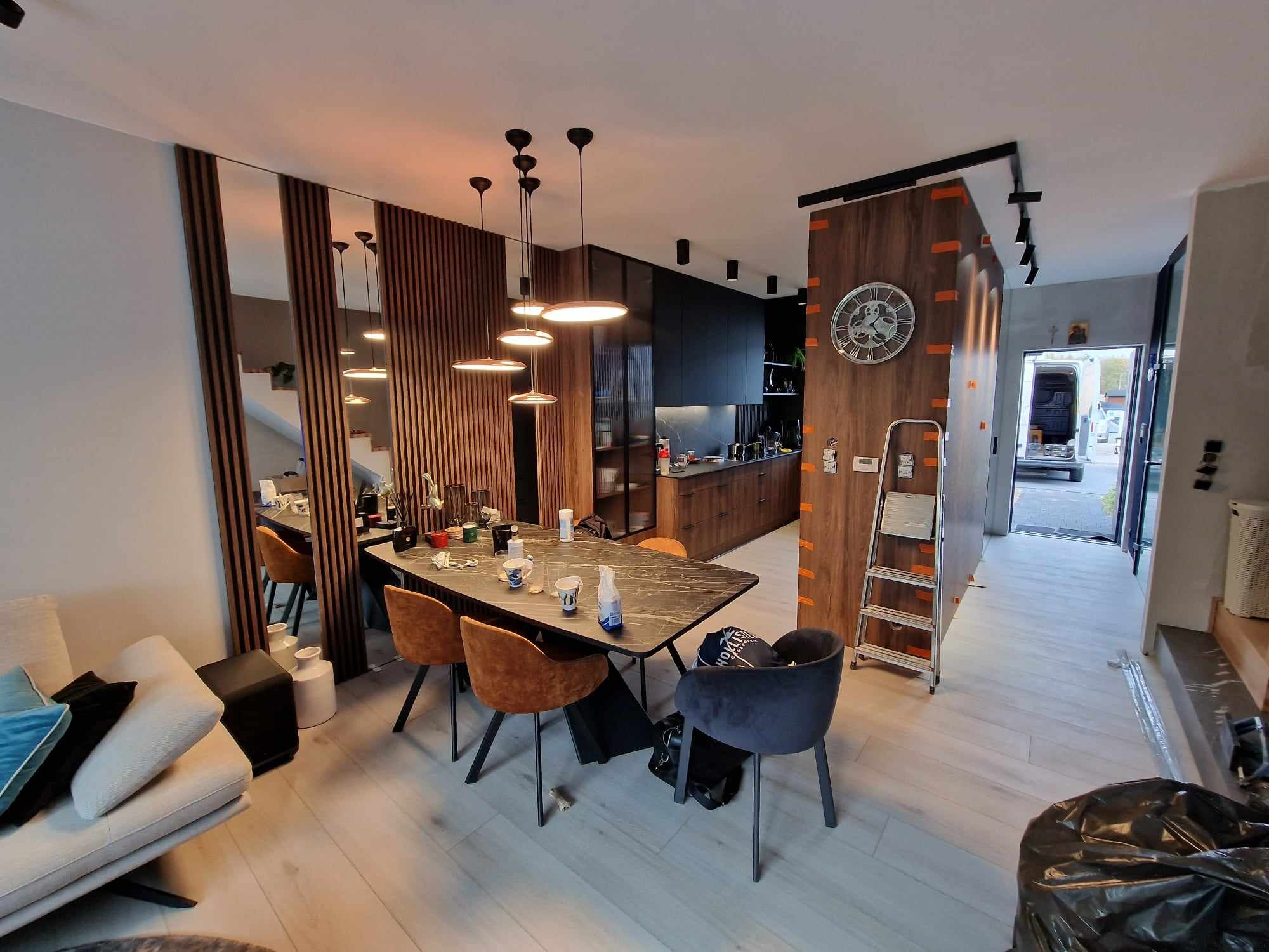 Nowoczesna kuchnia z ciemnymi szafkami i drewnianymi akcentami, widok na jadalnię z marmurowym stołem i krzesłami. Lustrzana ściana dodaje przestrzeni.