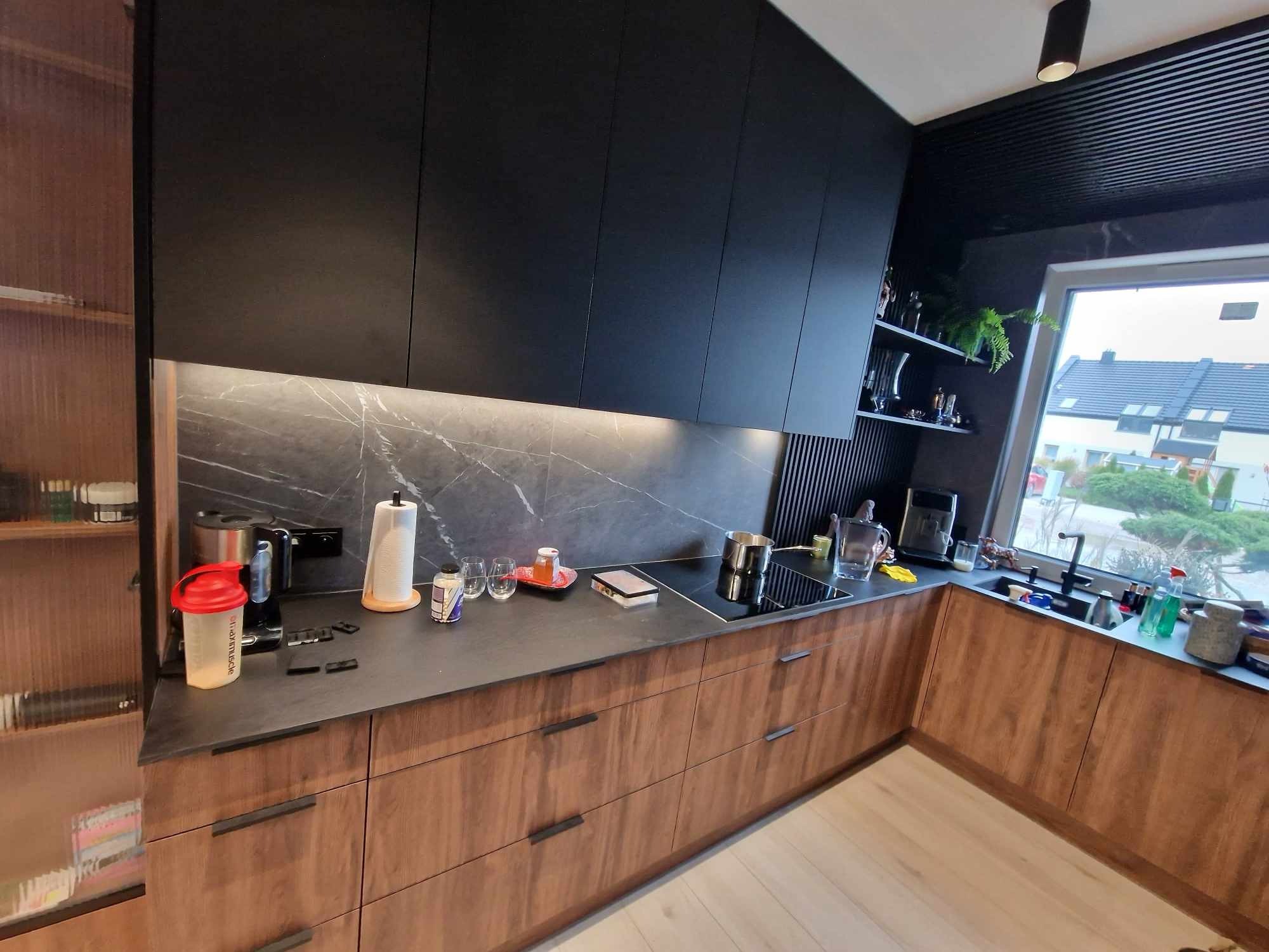 Nowoczesna kuchnia z drewnianymi frontami szafek i czarnymi szafkami górnymi, marmurowy backsplash, oświetlenie LED pod szafkami, widok na okno i elementy wyposażenia.