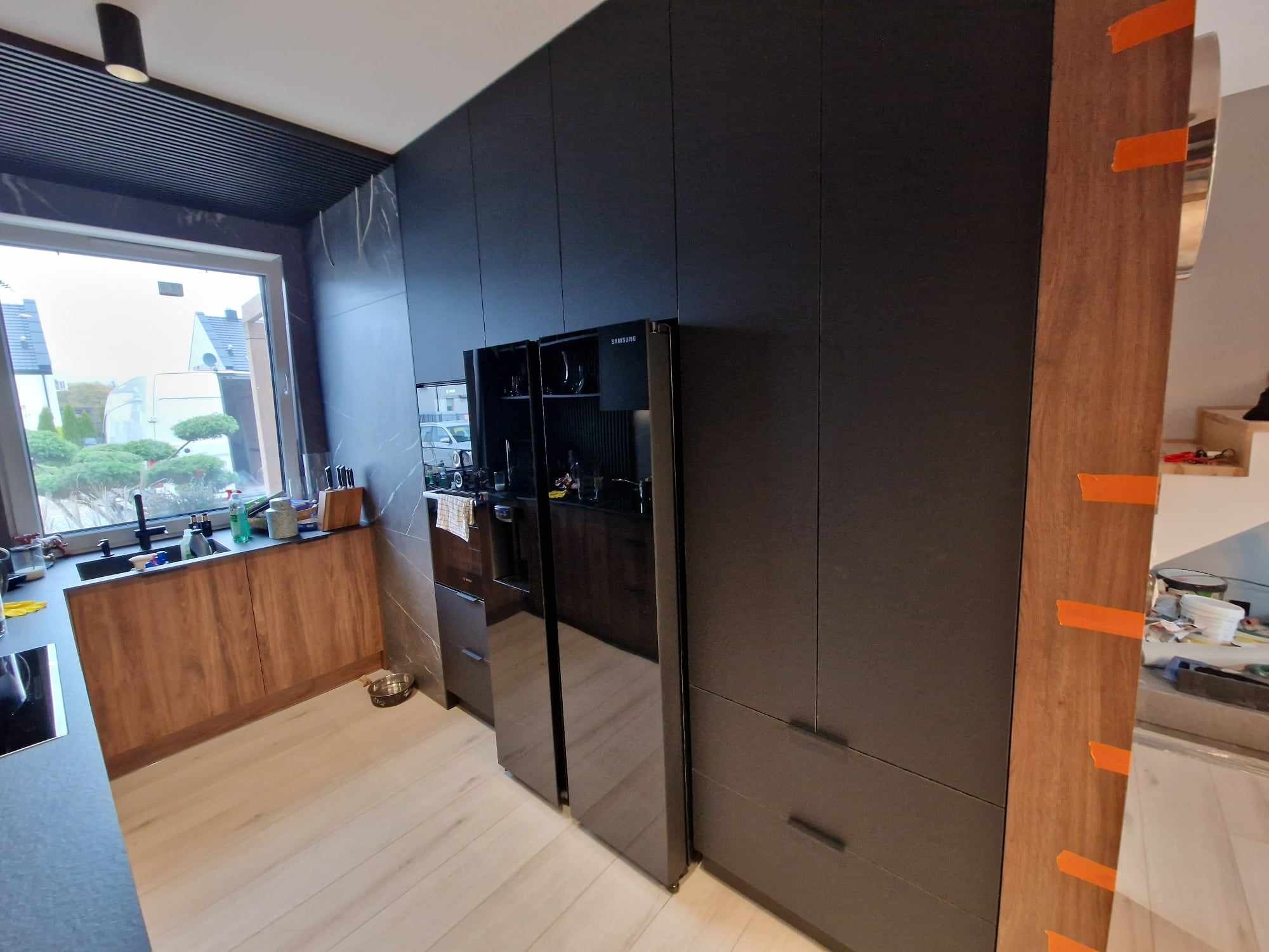 Nowoczesna kuchnia: zabudowa w odcieniach grafitu z dwoma lodówkami Samsung, drewniane szafki pod oknem i jasna podłoga. Widok na zewnątrz przez okno.