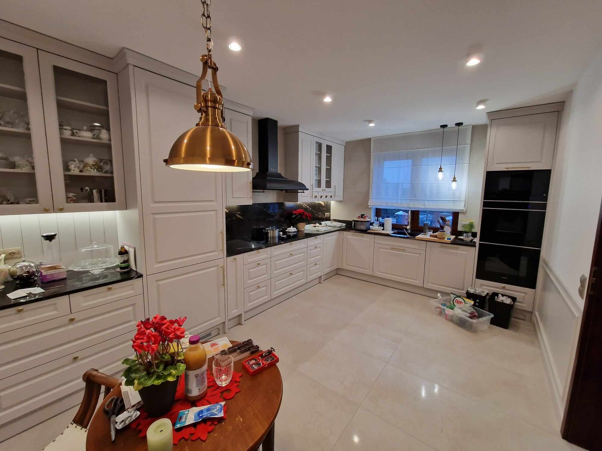 Jasna kuchnia w stylu klasycznym z białymi meblami i czarnym blatem. Nad stołem wisi mosiężna lampa. Widoczne detale: kwiaty, naczynia, okno z roletą.