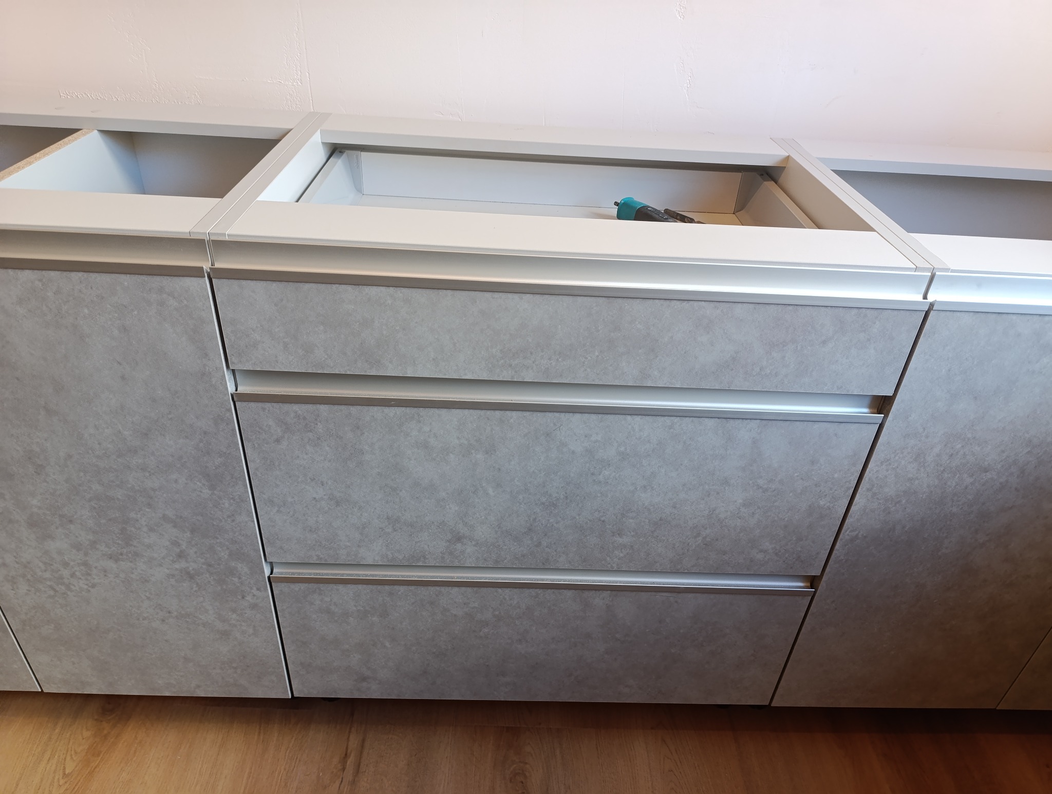 Szare, matowe fronty szuflad kuchennych z metalowymi uchwytami, widoczne puste przestrzenie szafek i narzędzie w środku. Ujęcie z dołu, ciepłe oświetlenie.