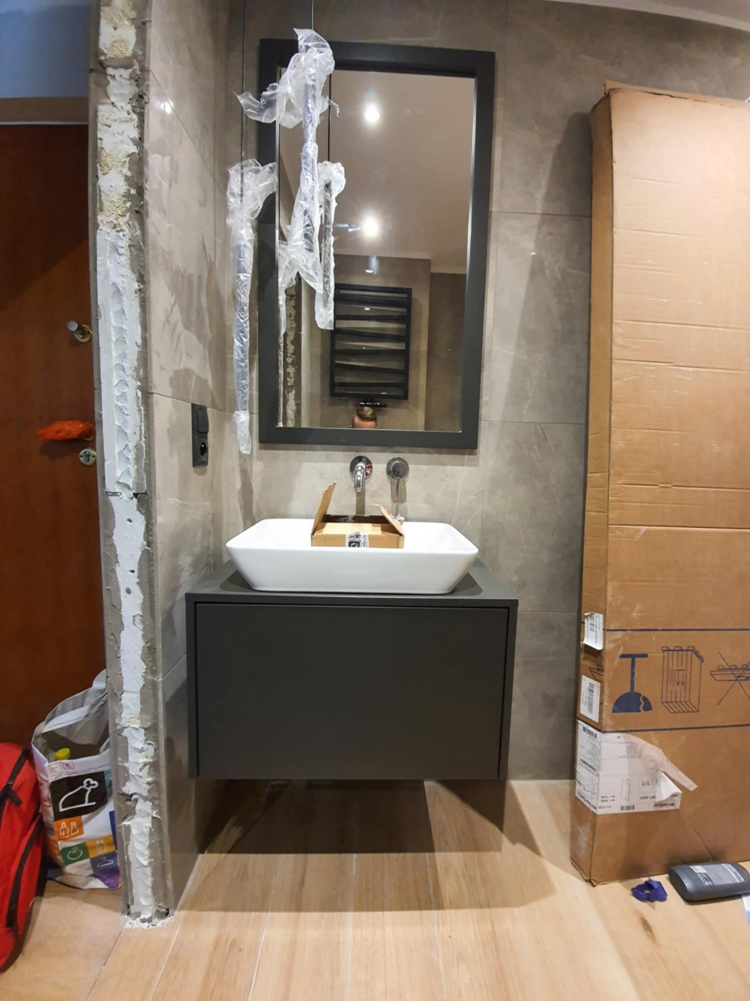 Montaż umywalki nablatowej z szarą szafką i lustrem w ramie. Widoczne niedokończone wykończenia ściany i kartonowe opakowanie obok. Ujęcie z bliska.