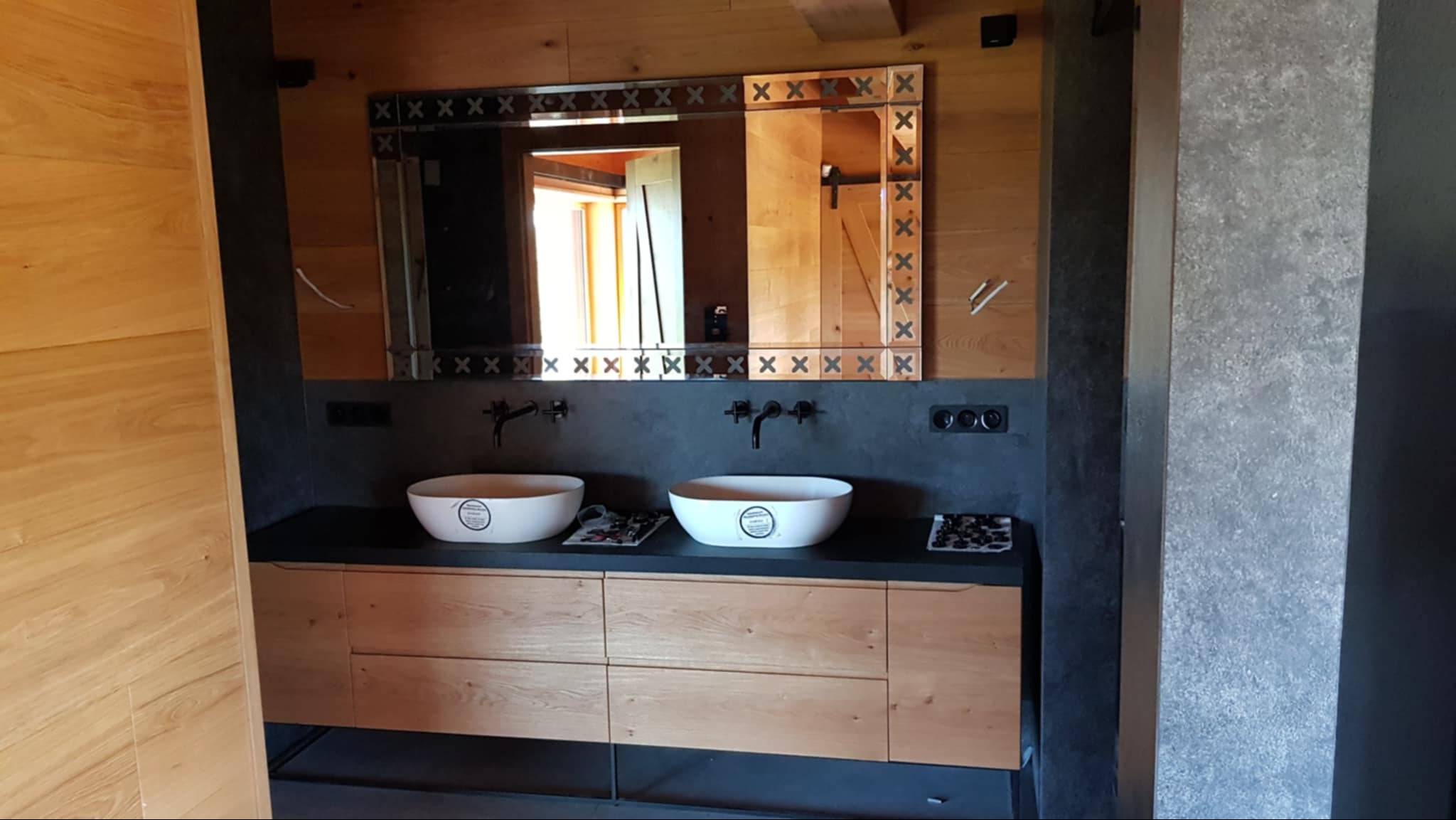 Drewniana szafka łazienkowa na wymiar z dwiema białymi umywalkami nablatowymi i lustrem w ramie z motywem krzyżyków, w łazience wykończonej drewnem i szarym betonem.