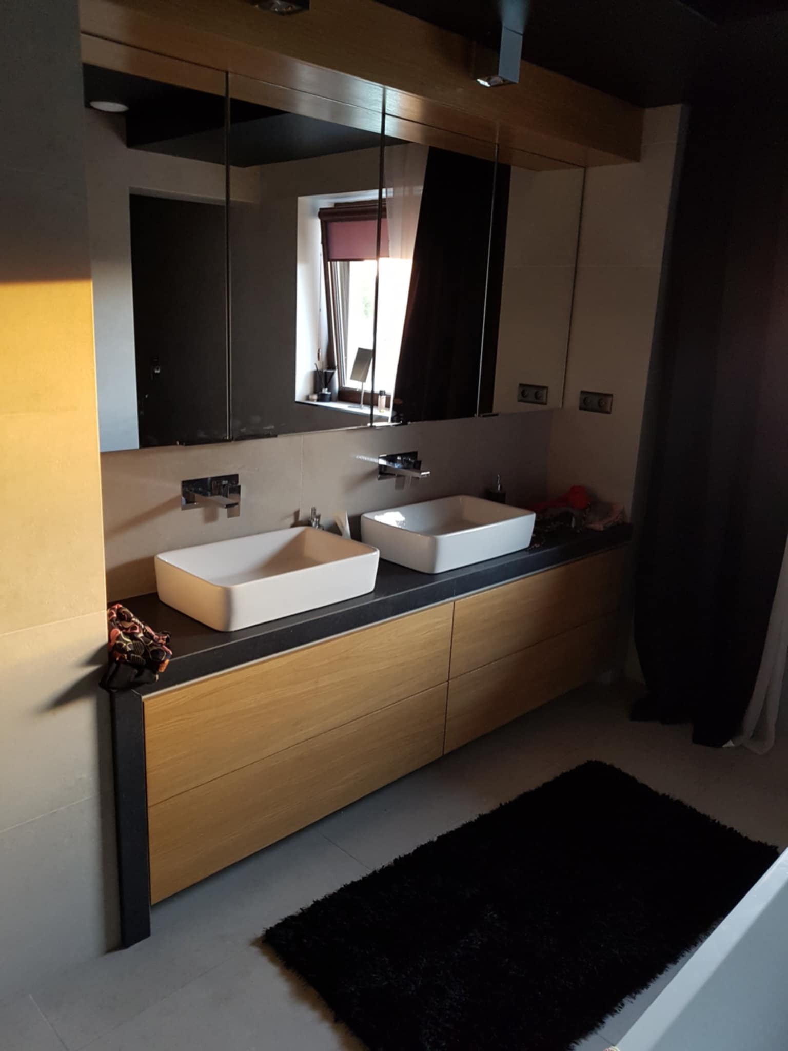 Nowoczesna łazienka z drewnianą szafką pod umywalki, czarnym blatem i dużym lustrem. Minimalistyczny design, stonowana kolorystyka, czarny dywanik.