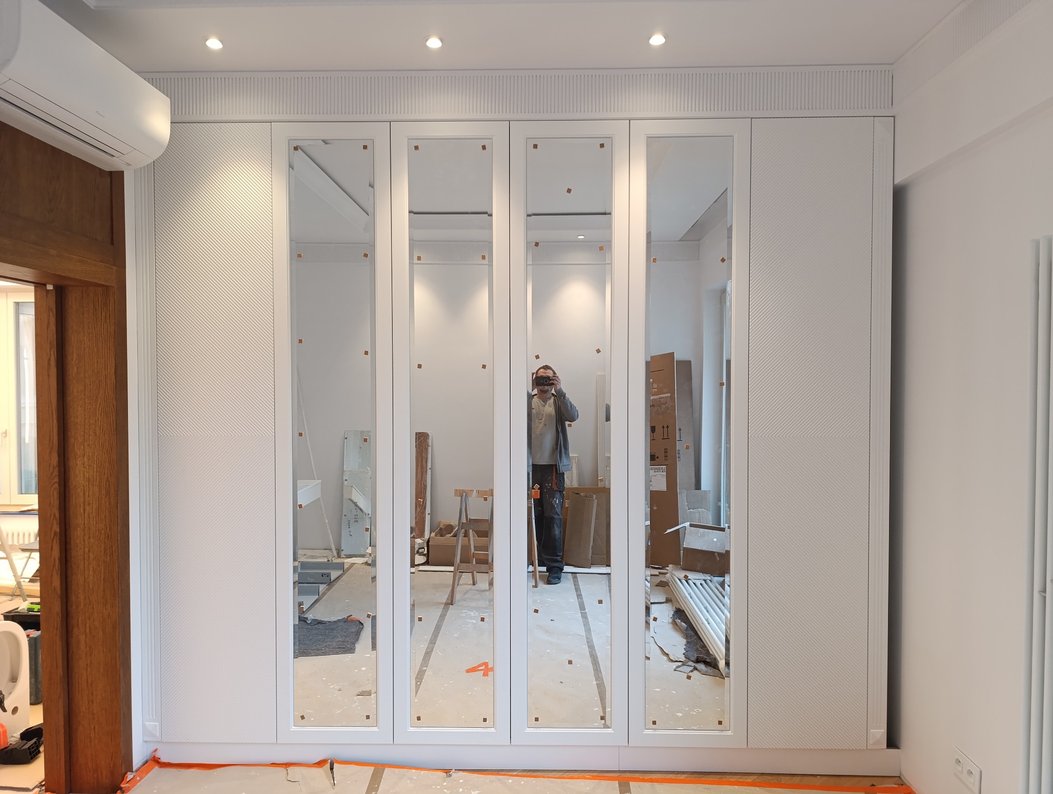 Biała szafa wnękowa z lustrzanymi drzwiami, widoczne zabezpieczenia na lustrach, w tle elementy remontu. Delikatna faktura na frontach szafy.