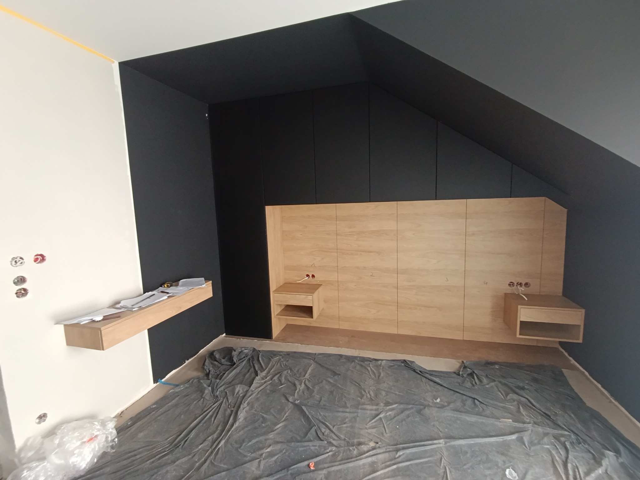 Montaż zabudowy w pokoju na poddaszu: czarne szafy wnękowe pod skosem, drewniane panele z szafkami nocnymi, podłoga zabezpieczona folią malarską.