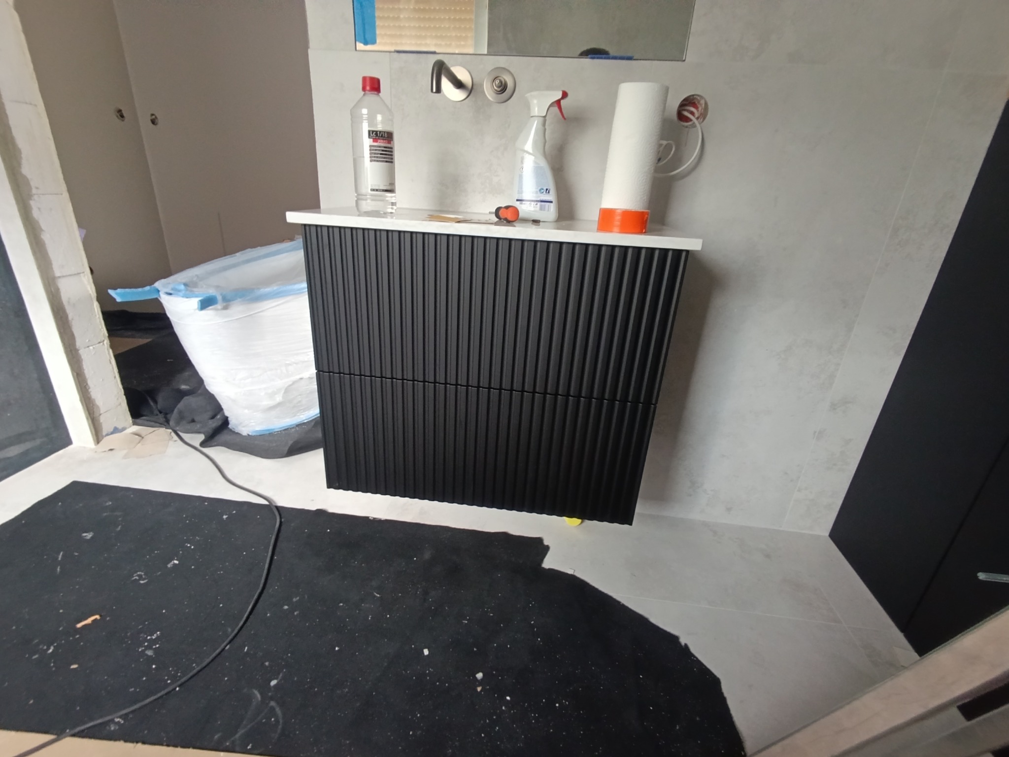 Czarna szafka łazienkowa z ryflowanym frontem, zamontowana w Śnieciskach. Na blacie chemia gospodarcza i narzędzia. W tle zabezpieczona folią wanna.