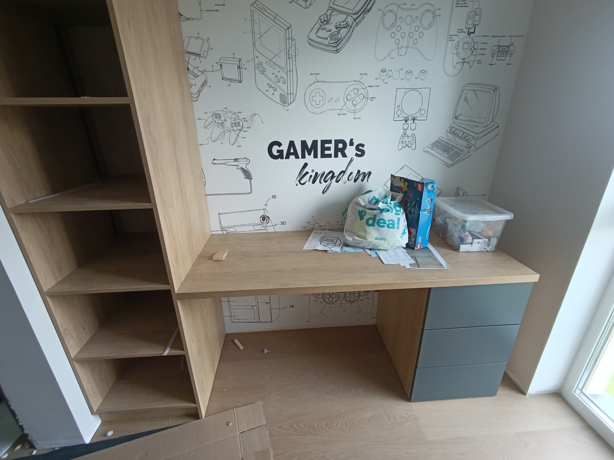 Biurko z szufladami i regał z półkami w pokoju gracza, z tapetą w motywie konsol i gier. Na blacie leżą przedmioty, widoczne jasne drewno i szare fronty szuflad.