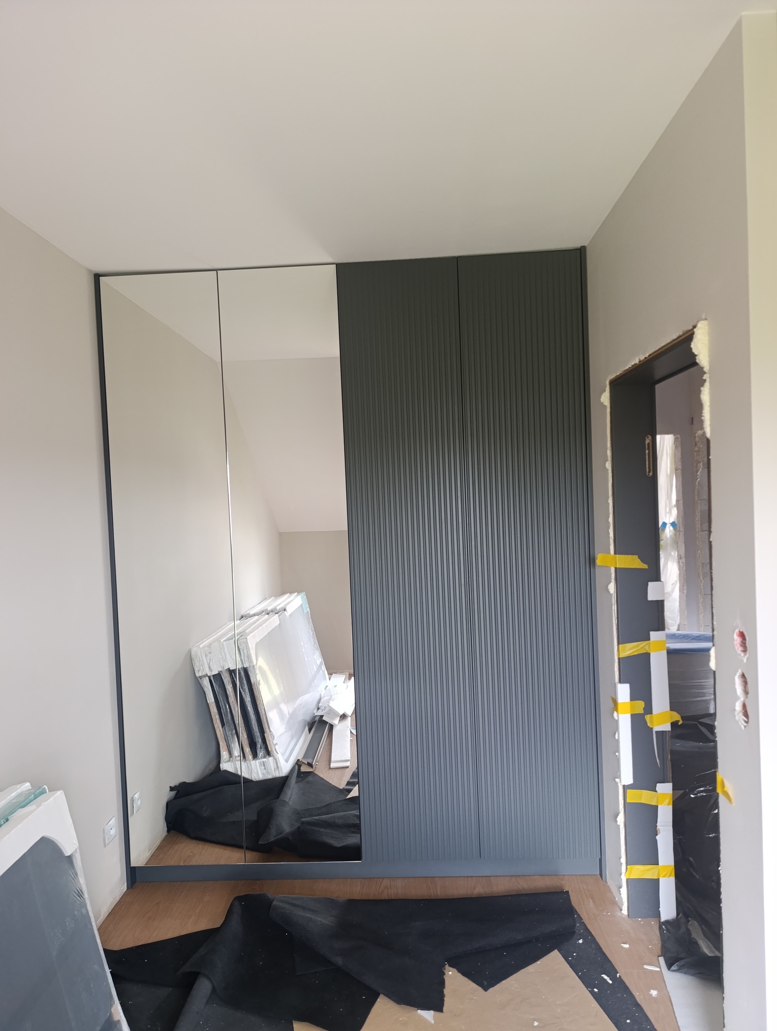 Szafa wnękowa z lustrzanymi drzwiami i ryflowanym frontem w szarym kolorze, widok na wnętrze szafy odbity w lustrze, zabezpieczona folią podłoga.