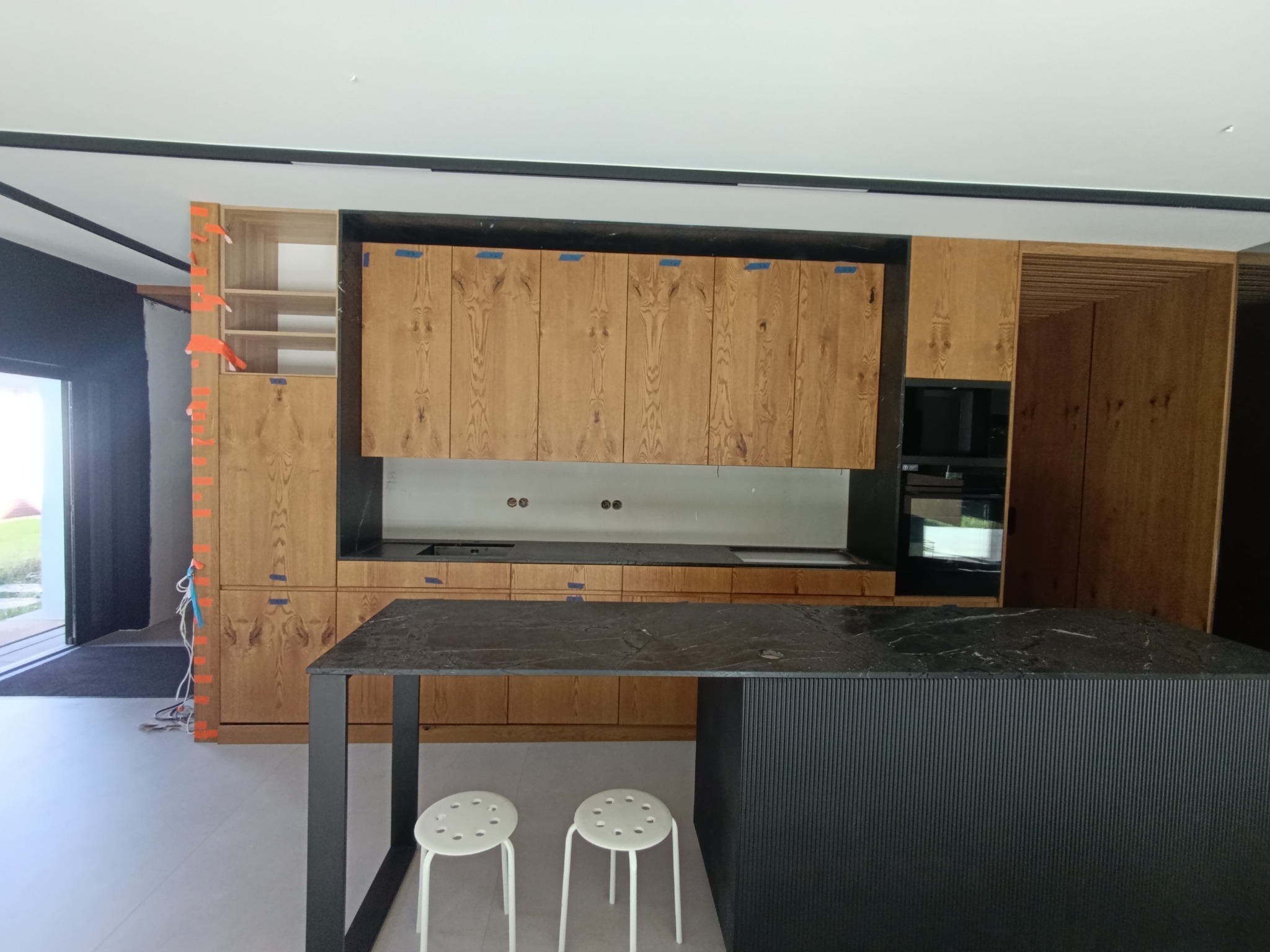 Nowoczesna kuchnia z drewnianymi frontami szafek, czarnym blatem i wyspą. Widoczne białe hokery i elementy wykończeniowe w trakcie montażu.