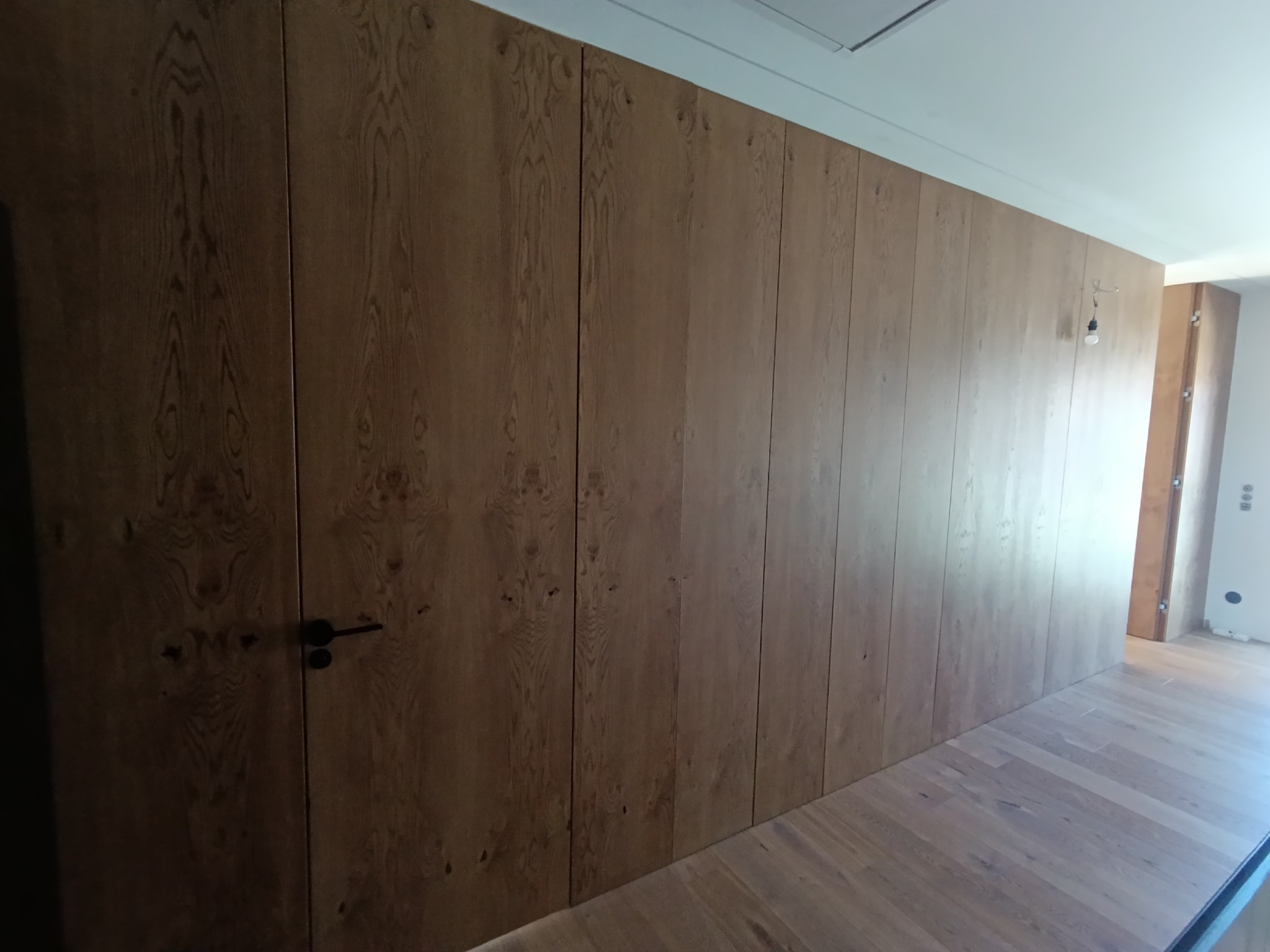 Długa, drewniana szafa wnękowa z ciemnymi uchwytami, dopasowana kolorystycznie do podłogi. Widok z perspektywy podłogi, ukazujący strukturę drewna i minimalistyczny design.