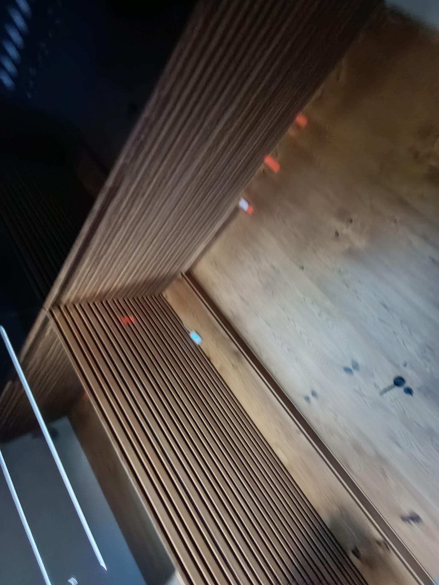 Drewniane lamele na ścianie, ułożone pod kątem, z widocznymi elementami montażowymi. Ciepłe oświetlenie podkreśla strukturę drewna. Nietypowy kadr.