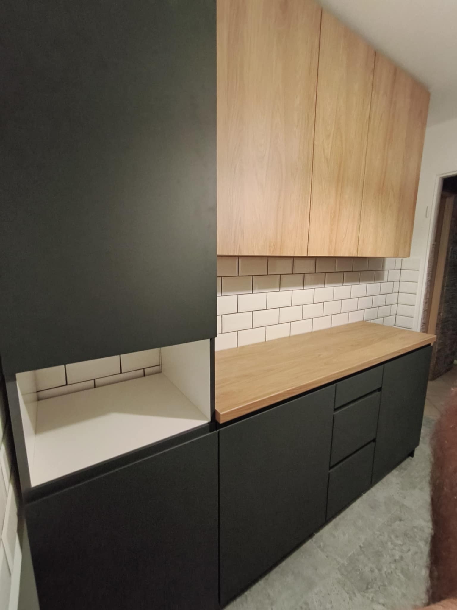 Nowoczesna kuchnia: czarne szafki dolne z drewnianym blatem i jasne szafki górne. Otwarta szafka z białym wnętrzem. Białe płytki na ścianie.