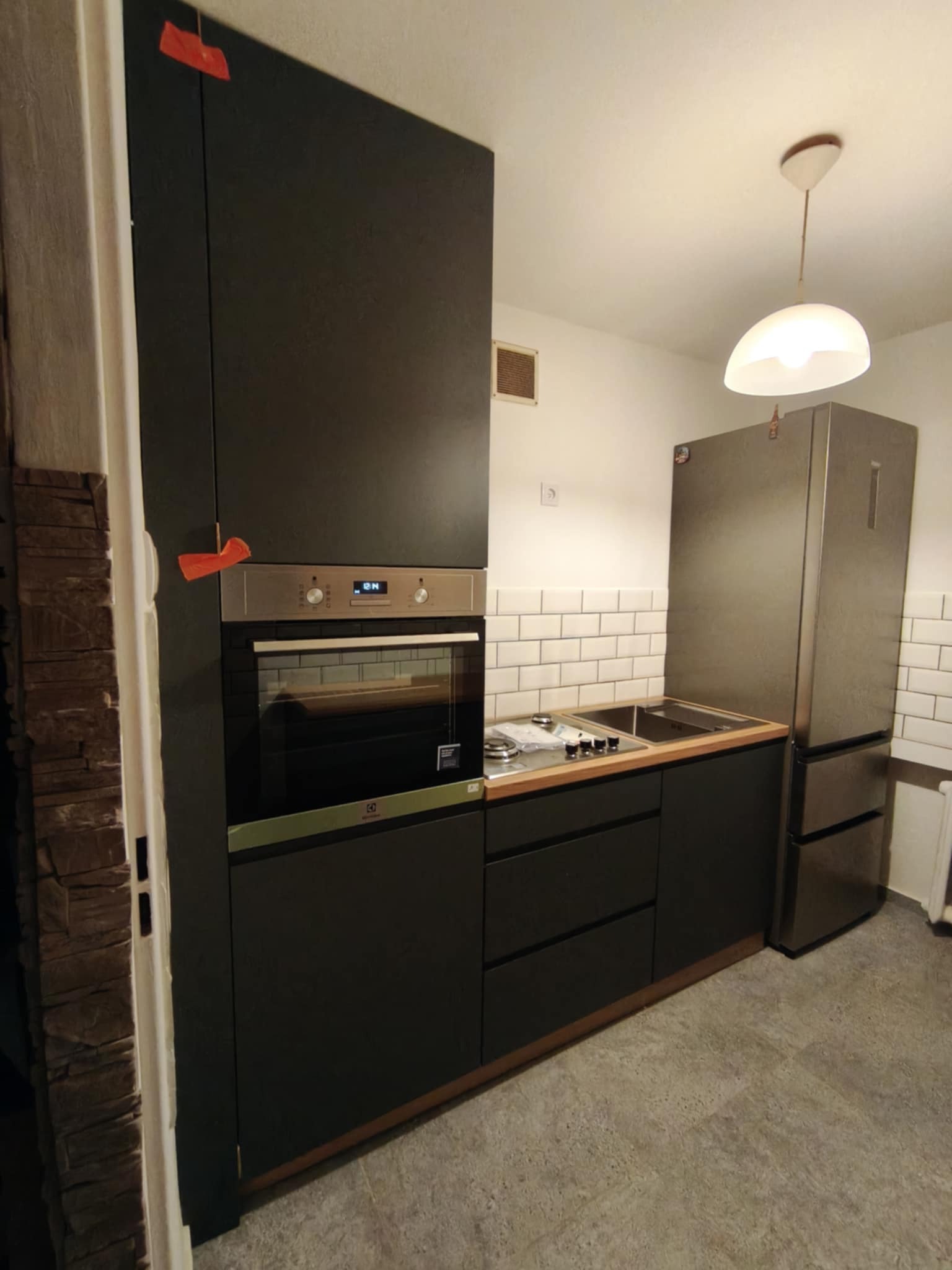 Ciemna zabudowa kuchenna z piekarnikiem i lodówką w małej kuchni, wykończona białymi płytkami i drewnianym blatem. Pomarańczowe elementy dekoracyjne na szafkach.