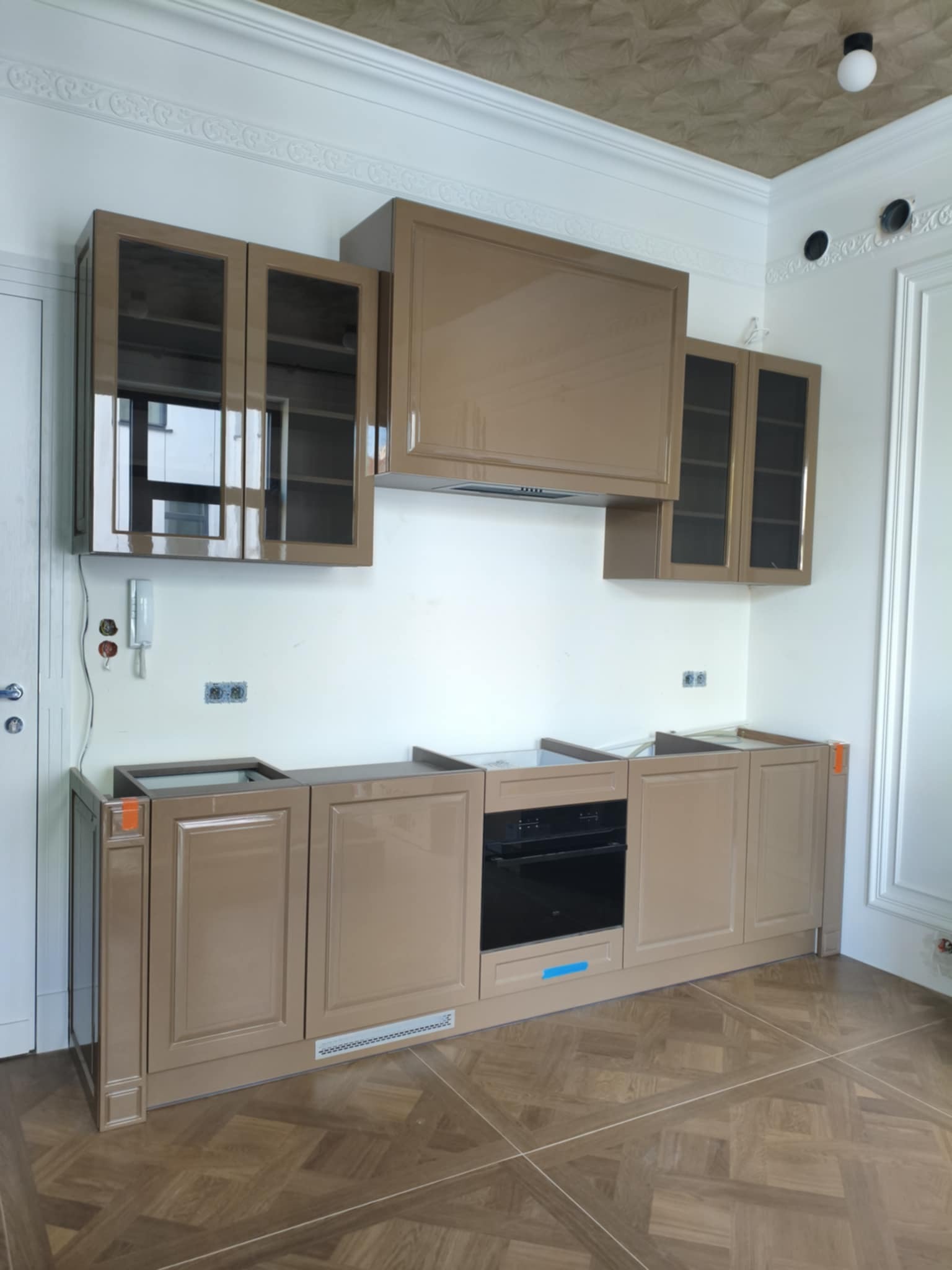 Montaż beżowej kuchni na wymiar z przeszklonymi szafkami wiszącymi i piekarnikiem w zabudowie. Widoczne niedokończone blaty i dekoracyjny sufit.