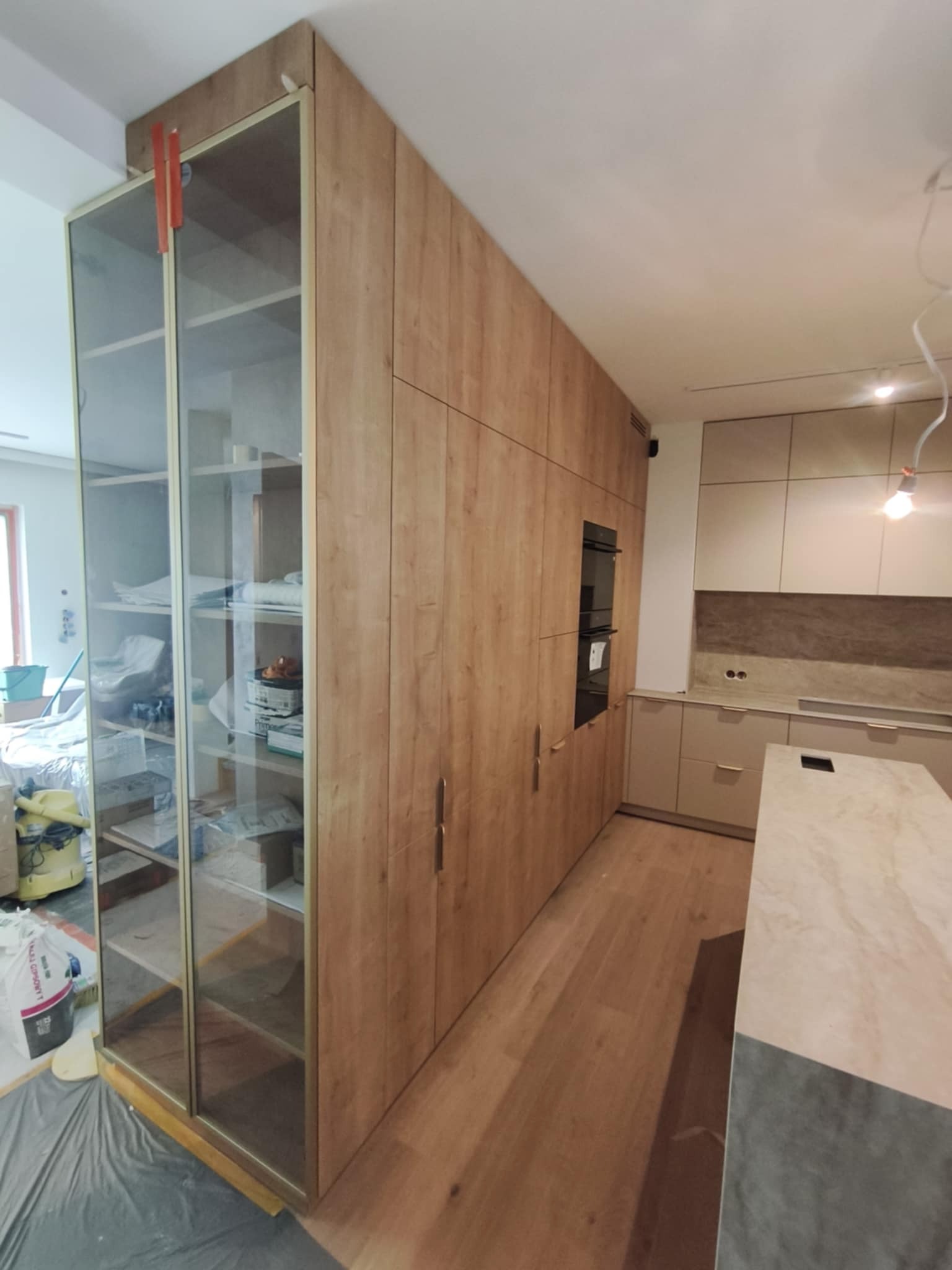 Jasna kuchnia z drewnianymi frontami szafek i przeszkloną witryną. Widoczne sprzęty AGD i blat. Podłoga drewniana. Częściowo zakryta folią ochronną.