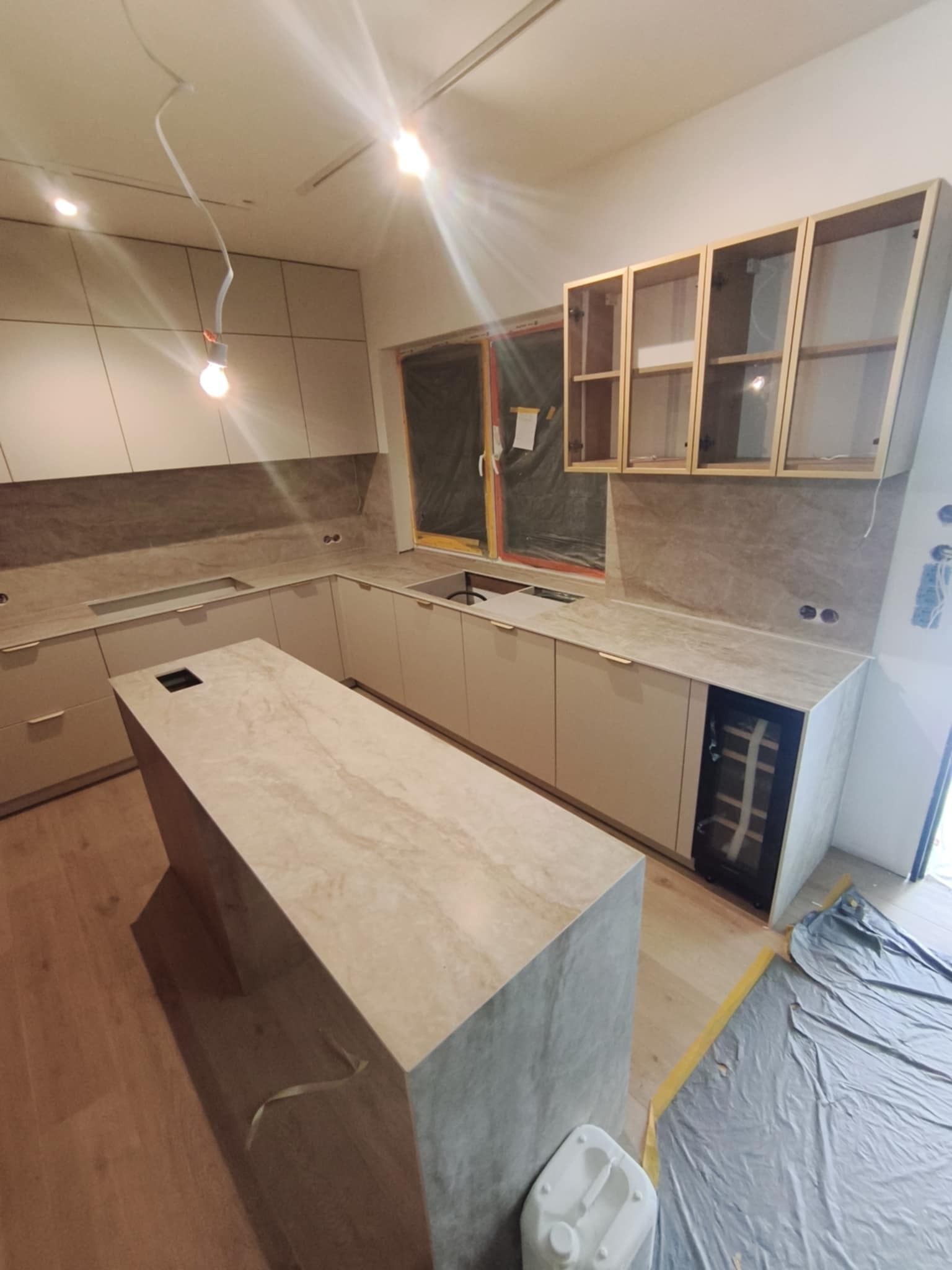 Jasna kuchnia z wyspą i przeszklonymi szafkami wiszącymi. Widoczne zabezpieczenia okna folią malarską i niedokończone wykończenie, sugerujące etap montażu.