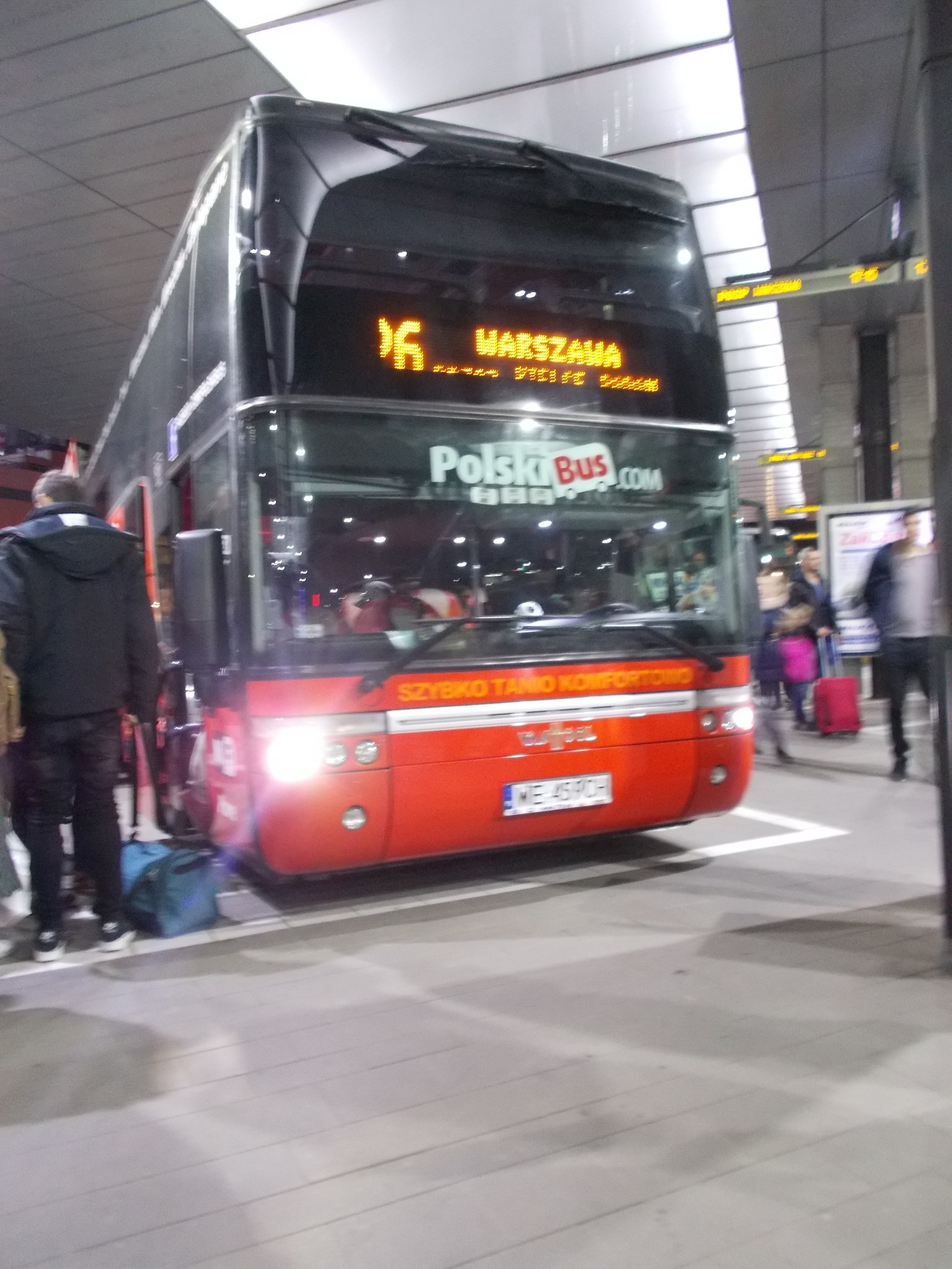 Czerwony autobus Polski Bus na dworcu, z wyświetlonym numerem linii 16 i kierunkiem Warszawa. Widoczni pasażerowie z bagażami i fragment peronu.