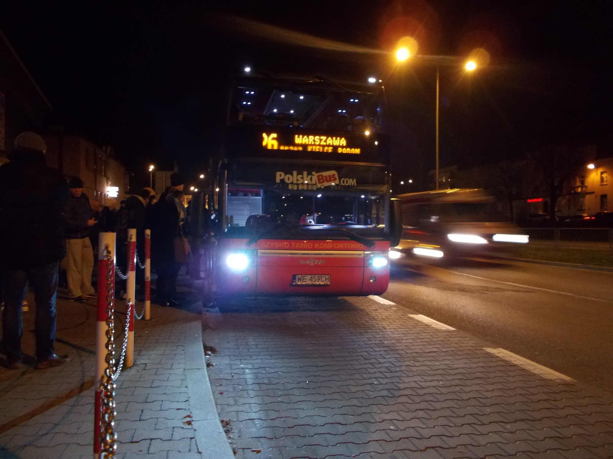 Autobus PolskiBus na przystanku nocą, z widocznymi pasażerami i światłami. Tablica z numerem linii i kierunkiem 'Warszawa' świeci jasno.