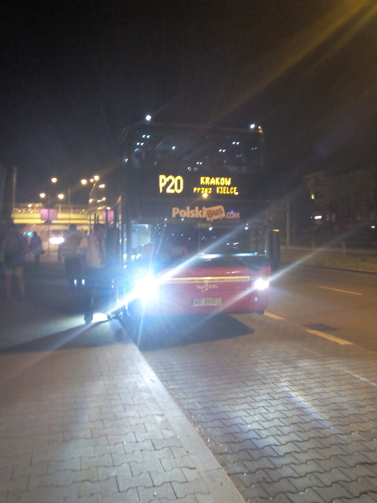 Nocny widok na dwupiętrowy autobus PolskiBus P20 z tablicą kierunkową Kraków/Kielce, oświetlony reflektorami, z pasażerami w tle na tle miejskich świateł.