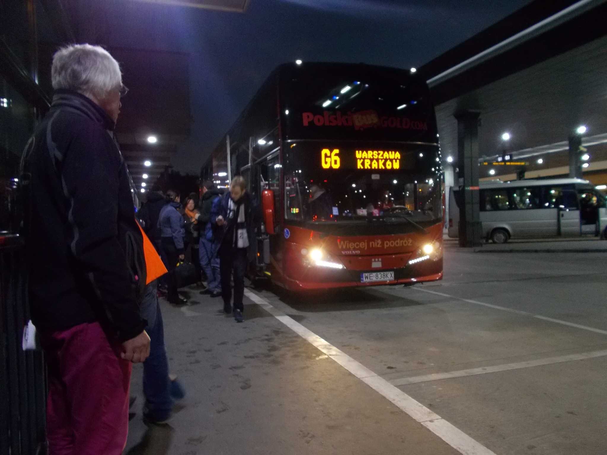 Czerwony autobus dalekobieżny PolskiBus G6 z podświetlonym napisem Warszawa-Kraków na dworcu autobusowym, pasażerowie wsiadają.