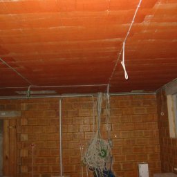FuseHill Mateusz Poncyleusz - Ceglana ściana i pomarańczowy sufit z widocznymi przewodami elektrycznymi w trakcie montażu. Widoczne kable zwisające ze ściany i sufitu.
