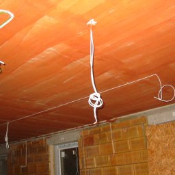 FuseHill Mateusz Poncyleusz - Instalacja elektryczna w surowym wnętrzu: przewody zwisają z pomarańczowego sufitu, widoczne okno i ściany z cegły oraz płyty OSB. Etap prac przed wykończeniem.