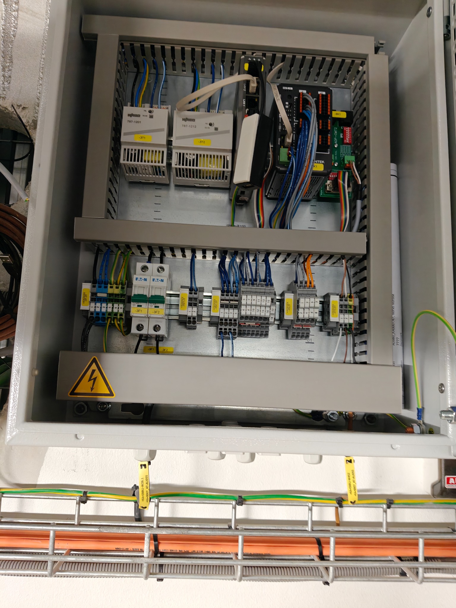 Otwarta szafa sterownicza z modułami, przewodami i zabezpieczeniami. Widoczny symbol ostrzegawczy i oznaczenia na przewodach. Instalacja w szarej obudowie.