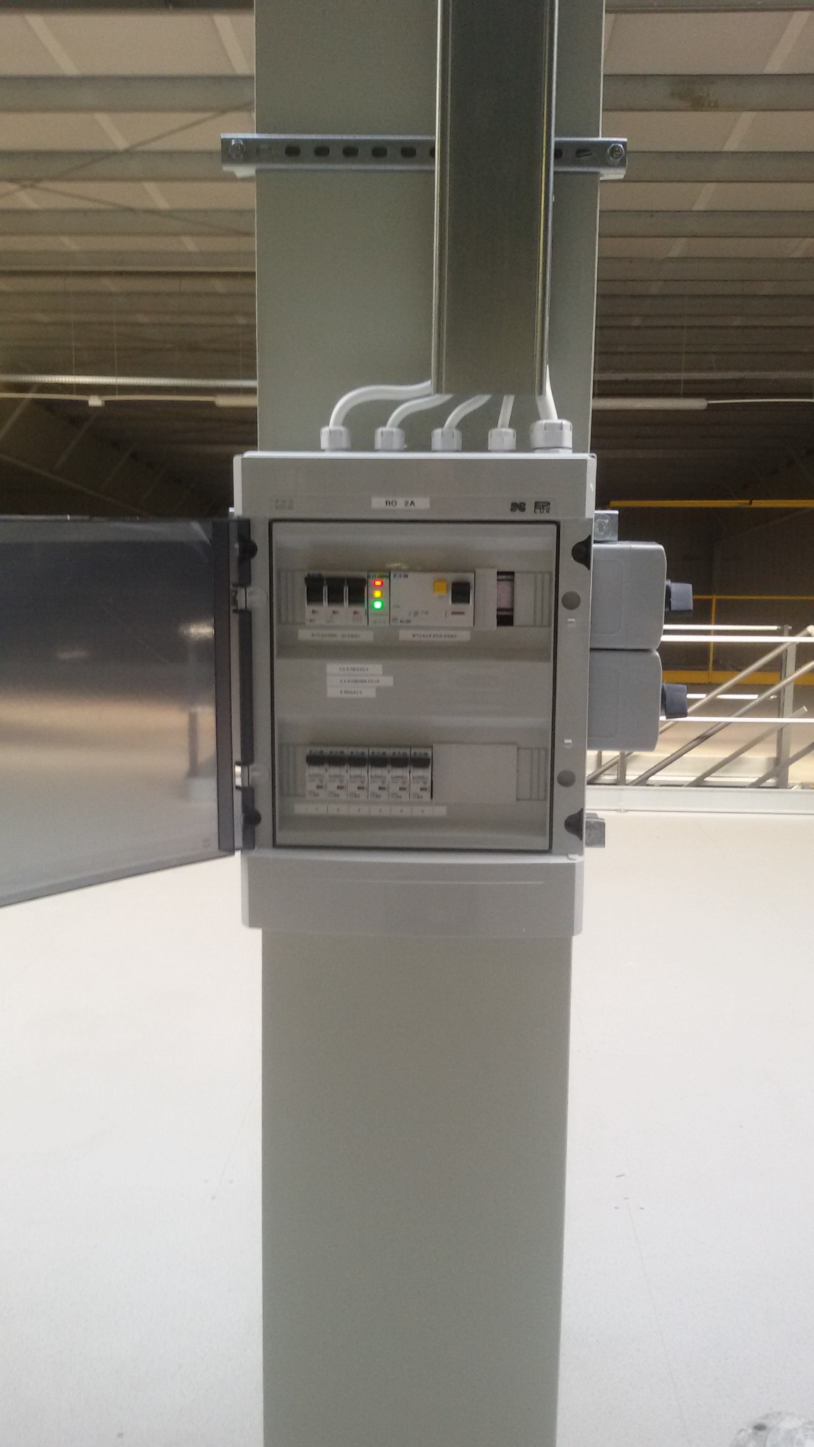 Otwarta szara skrzynka elektryczna z bezpiecznikami i przewodami, zamontowana na metalowym słupie w hali przemysłowej. Widoczne zielone diody sygnalizacyjne.