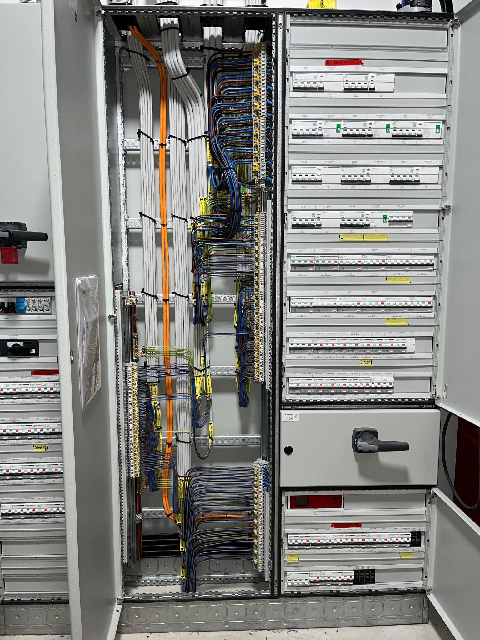 Otwarta szafa elektryczna z precyzyjnie ułożonymi kablami w różnych kolorach i rzędami bezpieczników. Widoczne etykiety i elementy sterujące.