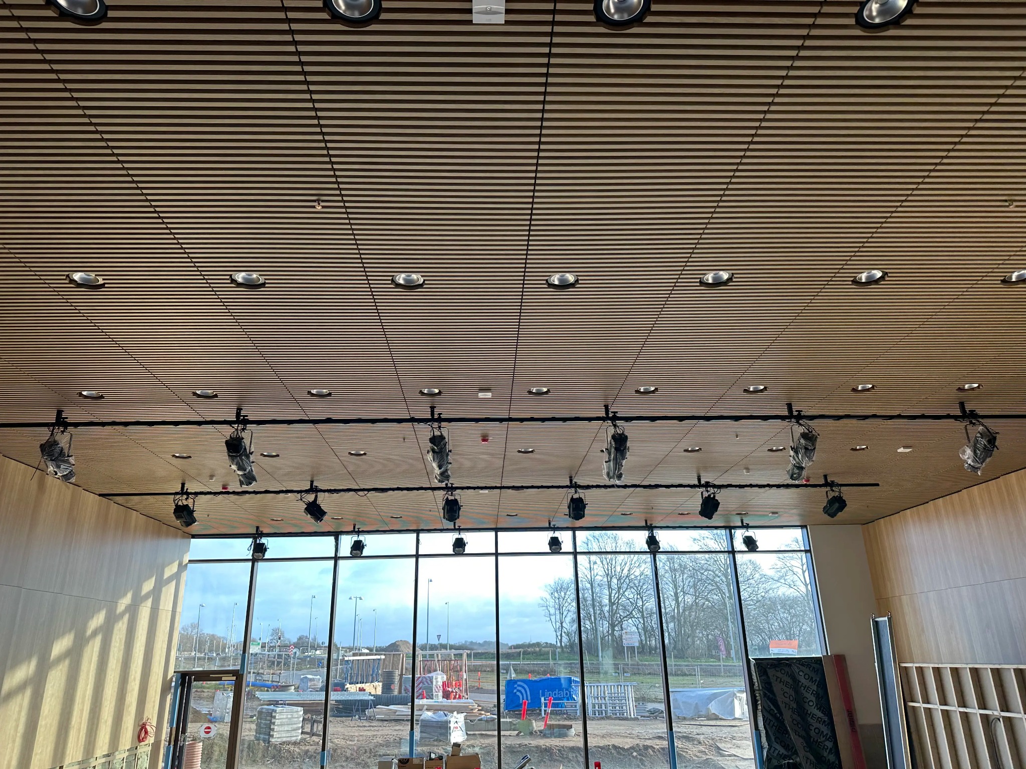 Drewniany sufit rastrowy z oświetleniem punktowym i reflektorami na szynach, widok z wnętrza budynku przez duże okna na plac budowy.