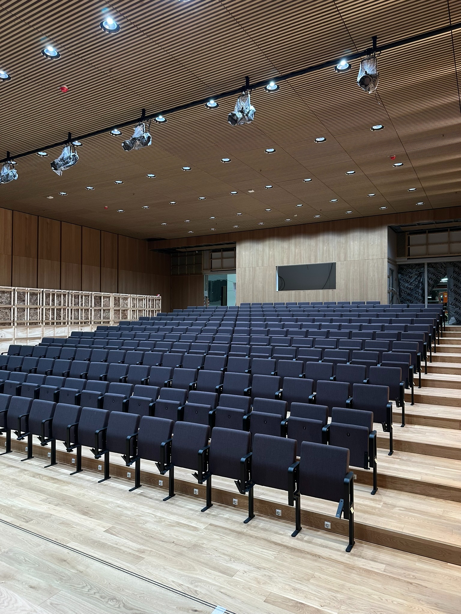 Sala widowiskowa z rzędami krzeseł na drewnianych stopniach, oświetlona reflektorami. Widoczne drewniane panele na ścianach i suficie. Część krzeseł jeszcze w folii.