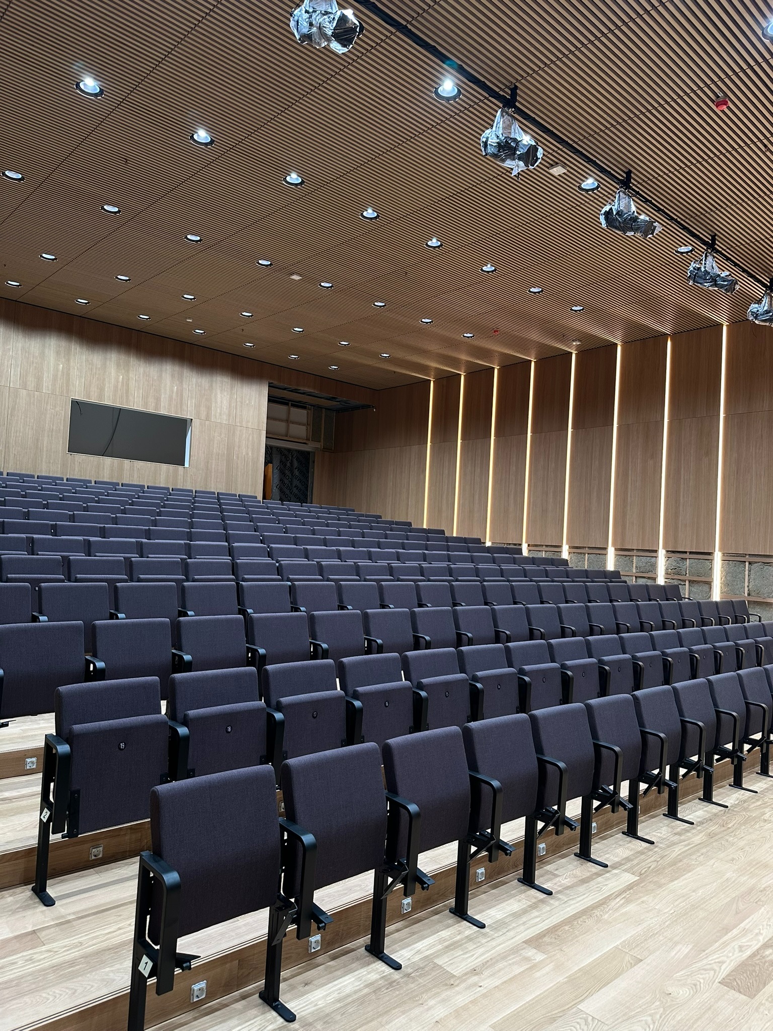 Sala konferencyjna z rzędami krzeseł na drewnianej podłodze, oświetlona punktowymi lampami i pionowymi panelami LED na ścianach. Widoczne projektory pod sufitem.